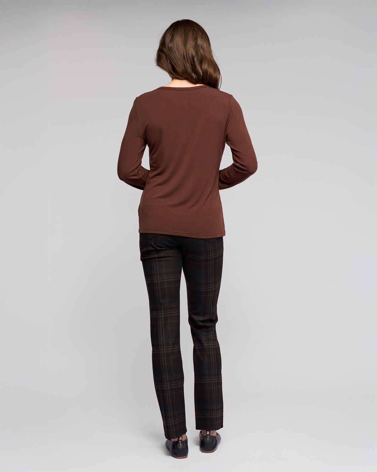 A woman faces away from the camera, wearing the Vassalli Layer Top in chocolate, relaxed-fit dark plaid pants, and black shoes, posed against a plain light gray background.
