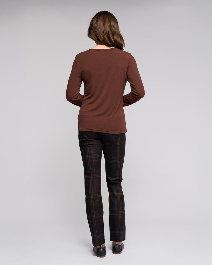 A woman faces away from the camera, wearing the Vassalli Layer Top in chocolate, relaxed-fit dark plaid pants, and black shoes, posed against a plain light gray background.
