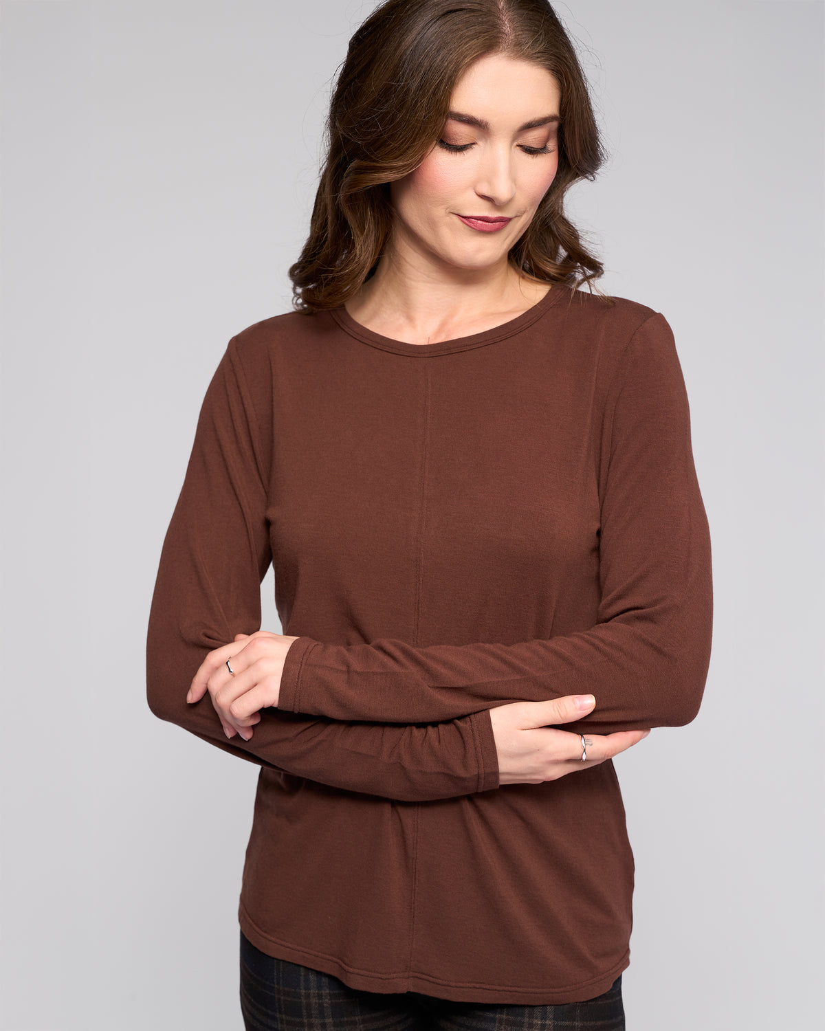 A woman with wavy brown hair wears the Vassalli Layer Top in chocolate—a relaxed fit, long-sleeve style—standing with arms crossed, smiling slightly and looking down against a plain light background.