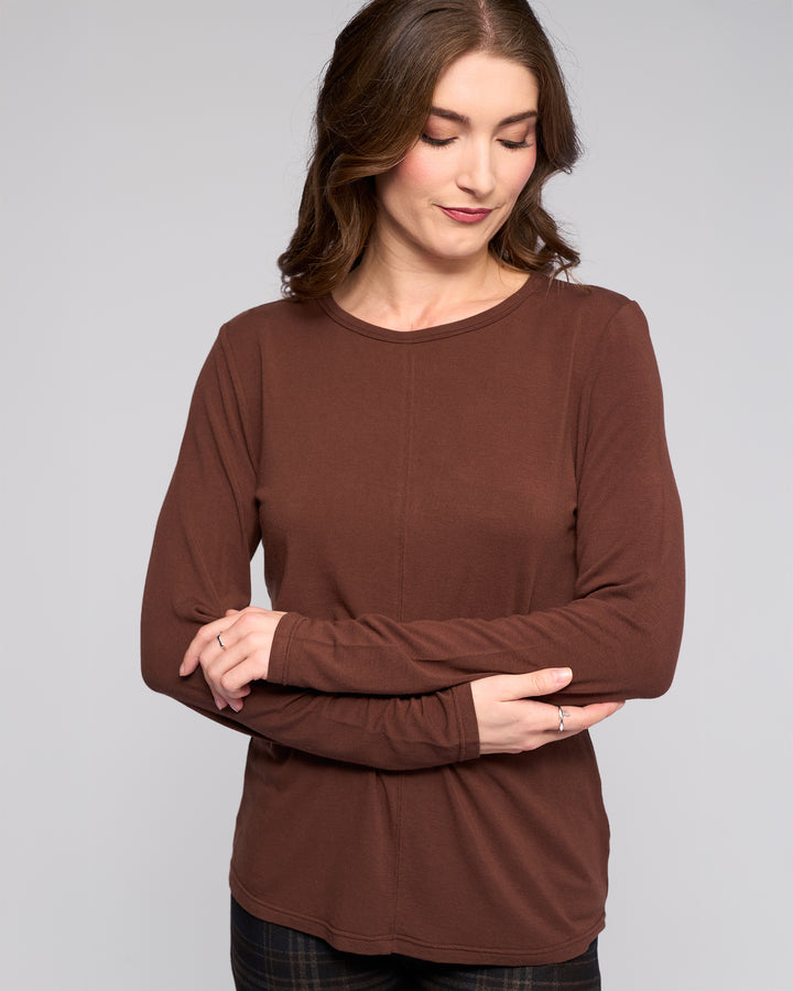 A woman with wavy brown hair wears the Vassalli Layer Top in chocolate—a relaxed fit, long-sleeve style—standing with arms crossed, smiling slightly and looking down against a plain light background.