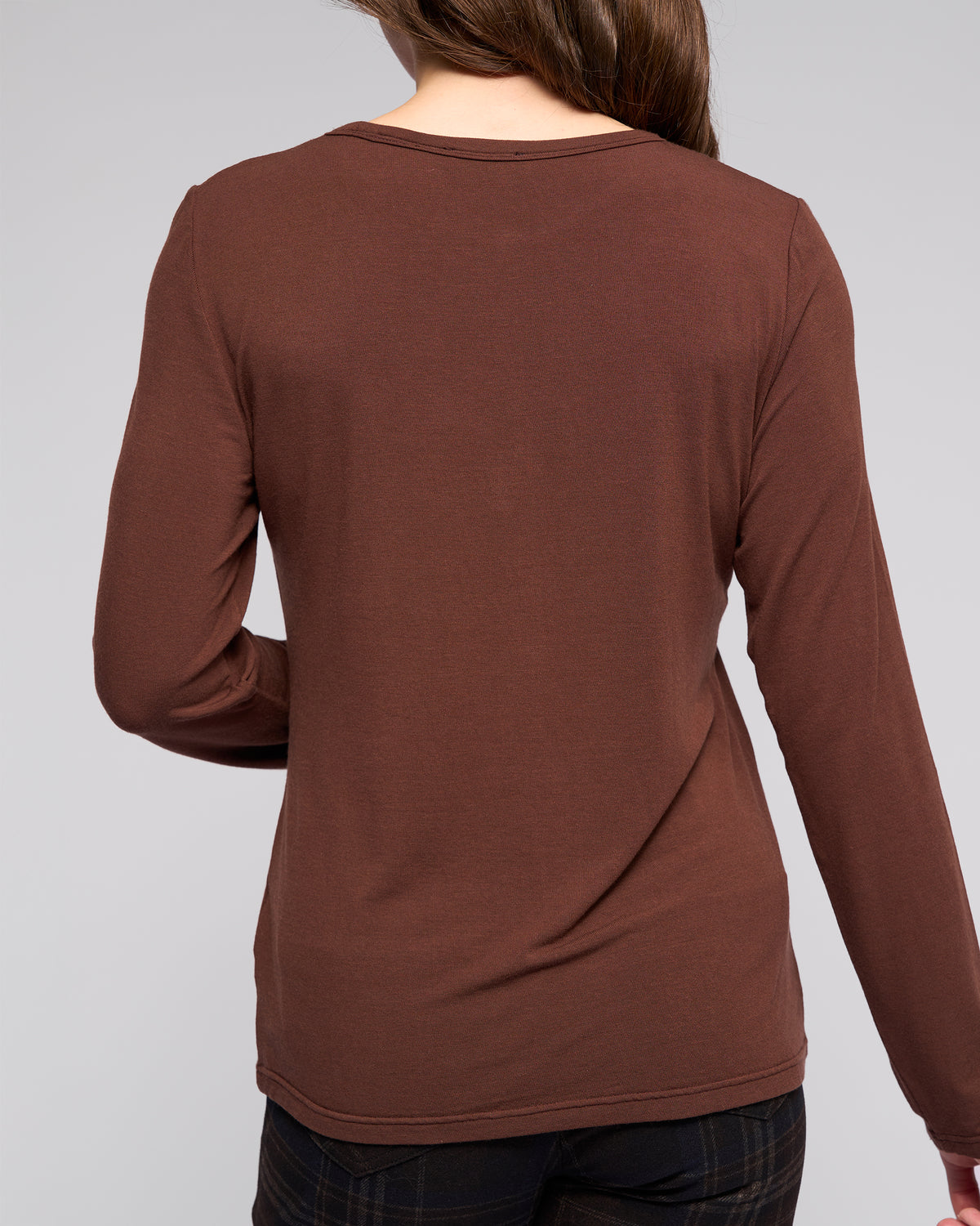 A woman with long brown hair, seen from the back, wears the Vassalli Layer Top - Chocolate in a relaxed fit with dark plaid pants against a plain light gray background.