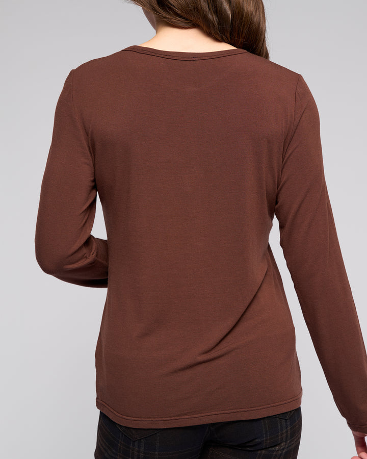 A woman with long brown hair, seen from the back, wears the Vassalli Layer Top - Chocolate in a relaxed fit with dark plaid pants against a plain light gray background.