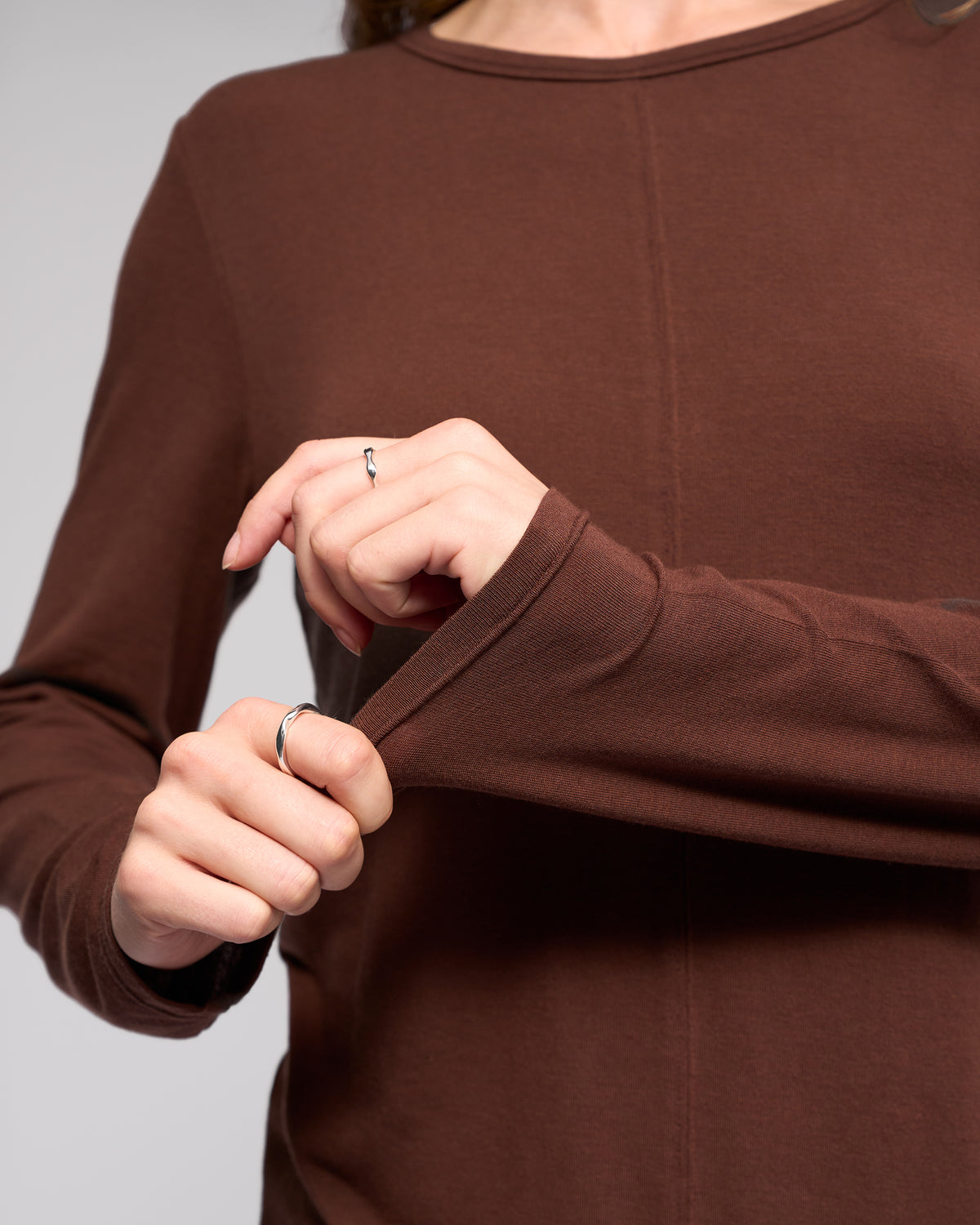 Close-up of a person wearing the Vassalli Layer Top in chocolate, relaxed fit. Only the torso and hands are visible, with two simple rings—one on each hand—as they tug at the sleeve.
