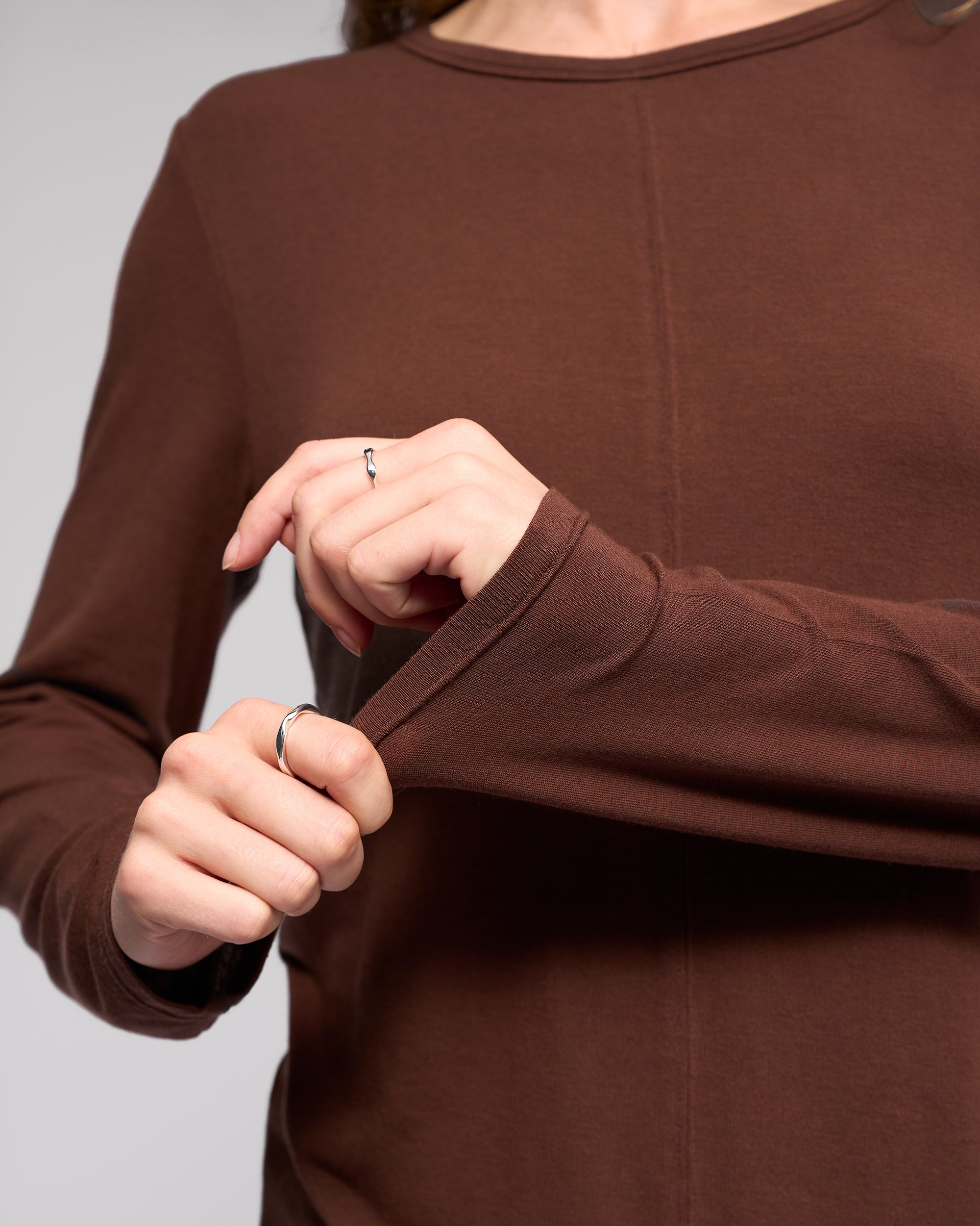 Close-up of a person wearing the Vassalli Layer Top in chocolate, relaxed fit. Only the torso and hands are visible, with two simple rings—one on each hand—as they tug at the sleeve.