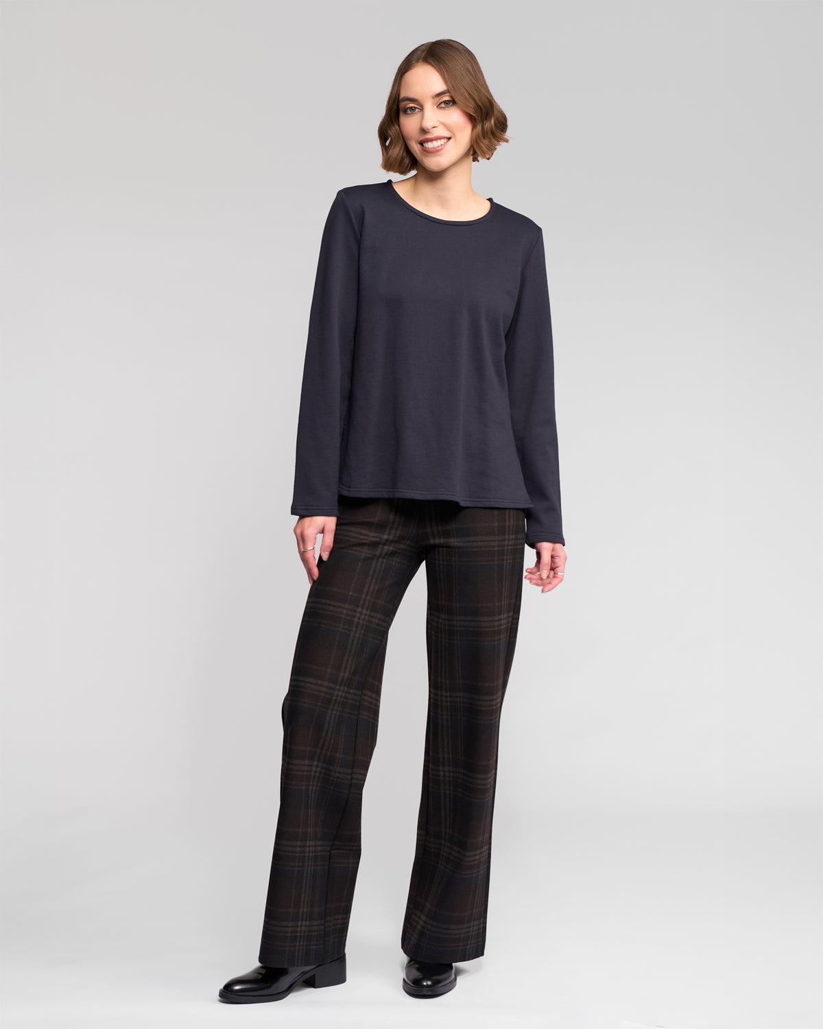 A woman with short brown hair is smiling against a light gray background, wearing the Vassalli Chase Top in Ink, paired with plaid wide-leg pants and black shoes for a comfortable yet stylish look.