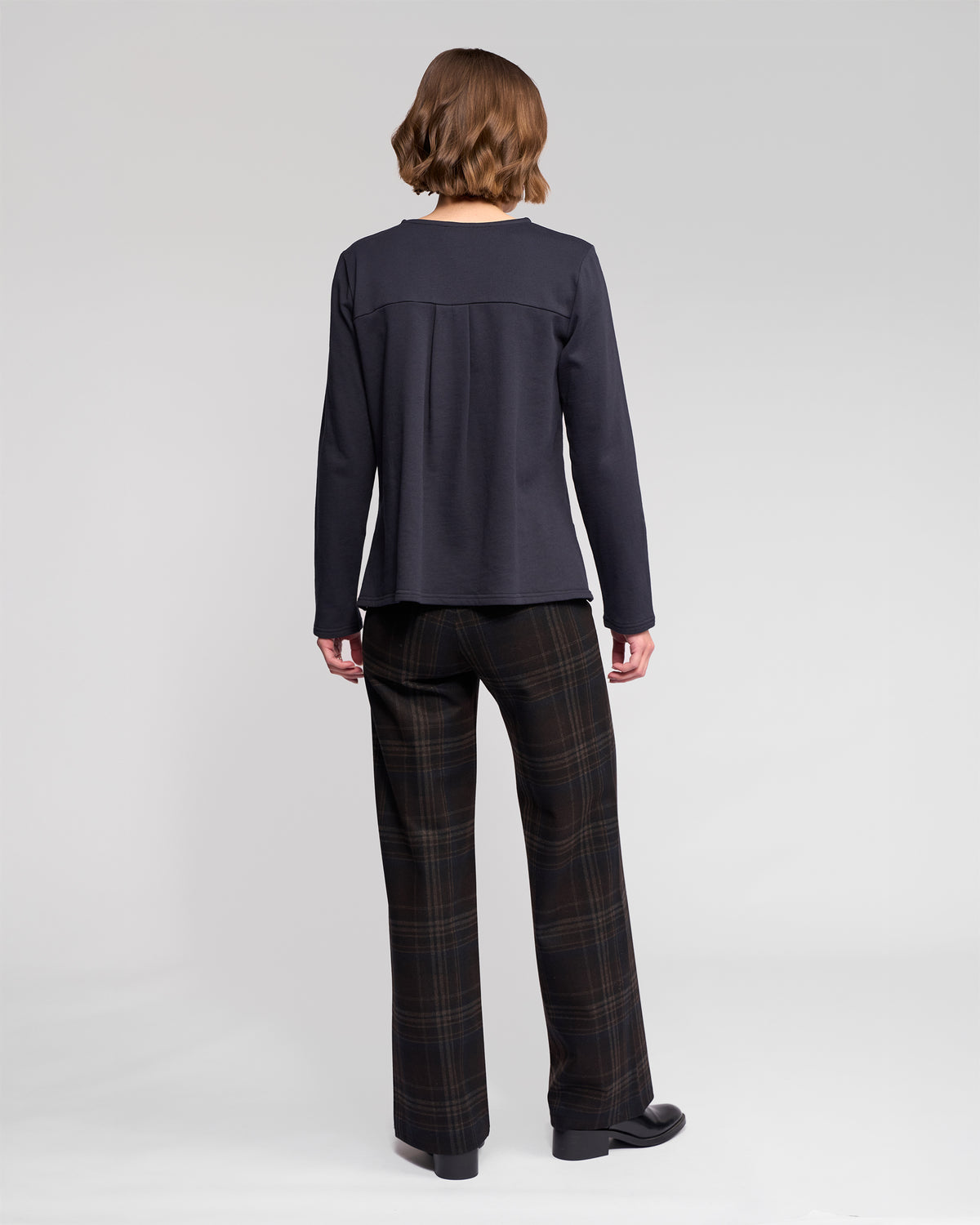 A person with short, wavy brown hair stands facing away from the camera, wearing the Vassalli Chase Top - Ink and loose dark plaid pants against a plain light background.