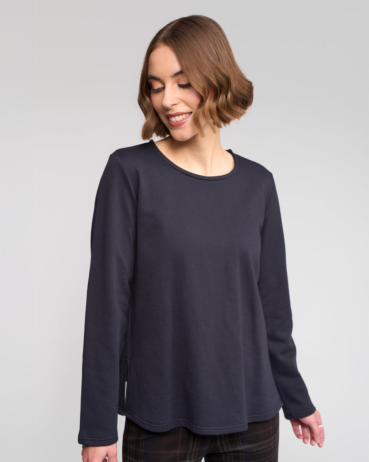 A woman with short, wavy brown hair is smiling and looking down while wearing the Vassalli Chase Top in Ink, a relaxed-fit long sleeve top, and standing against a plain light background.