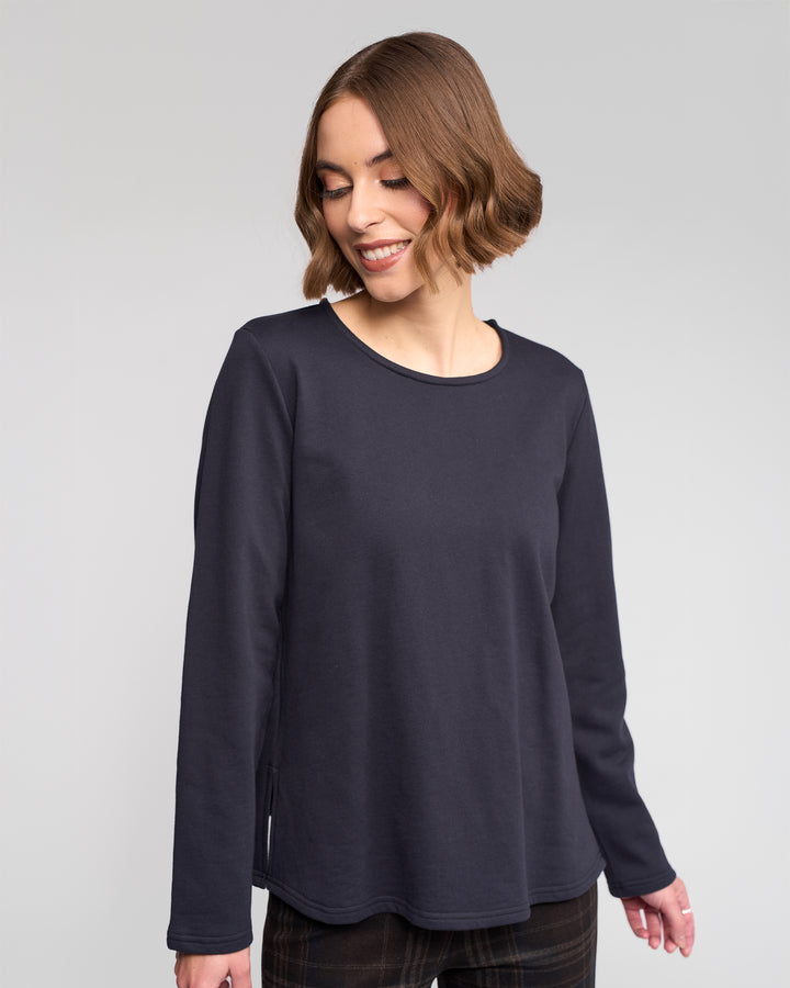 A woman with short, wavy brown hair is smiling and looking down while wearing the Vassalli Chase Top in Ink, a relaxed-fit long sleeve top, and standing against a plain light background.