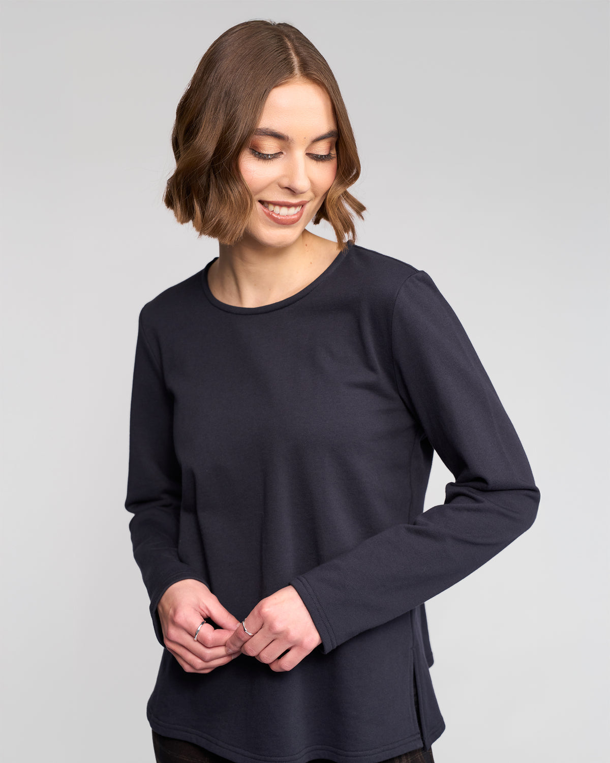 A woman with short brown hair smiles while looking down, wearing the Vassalli Chase Top in Ink. She stands against a plain light gray background, her hands gently touching in front of her.
