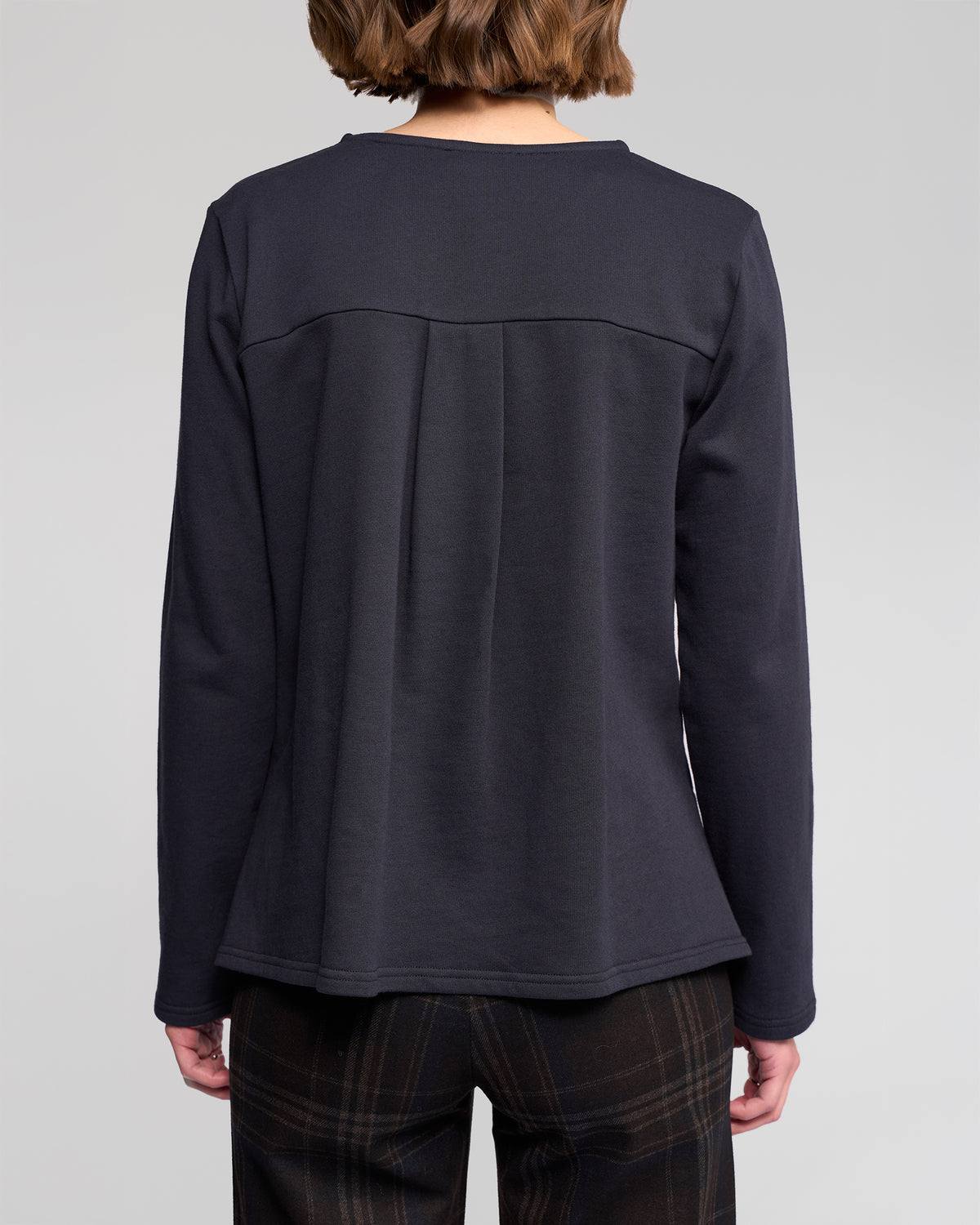 A person with short brown hair, seen from behind, is wearing the Vassalli Chase Top in Ink and dark plaid winter-weight cotton pants against a plain light gray background.