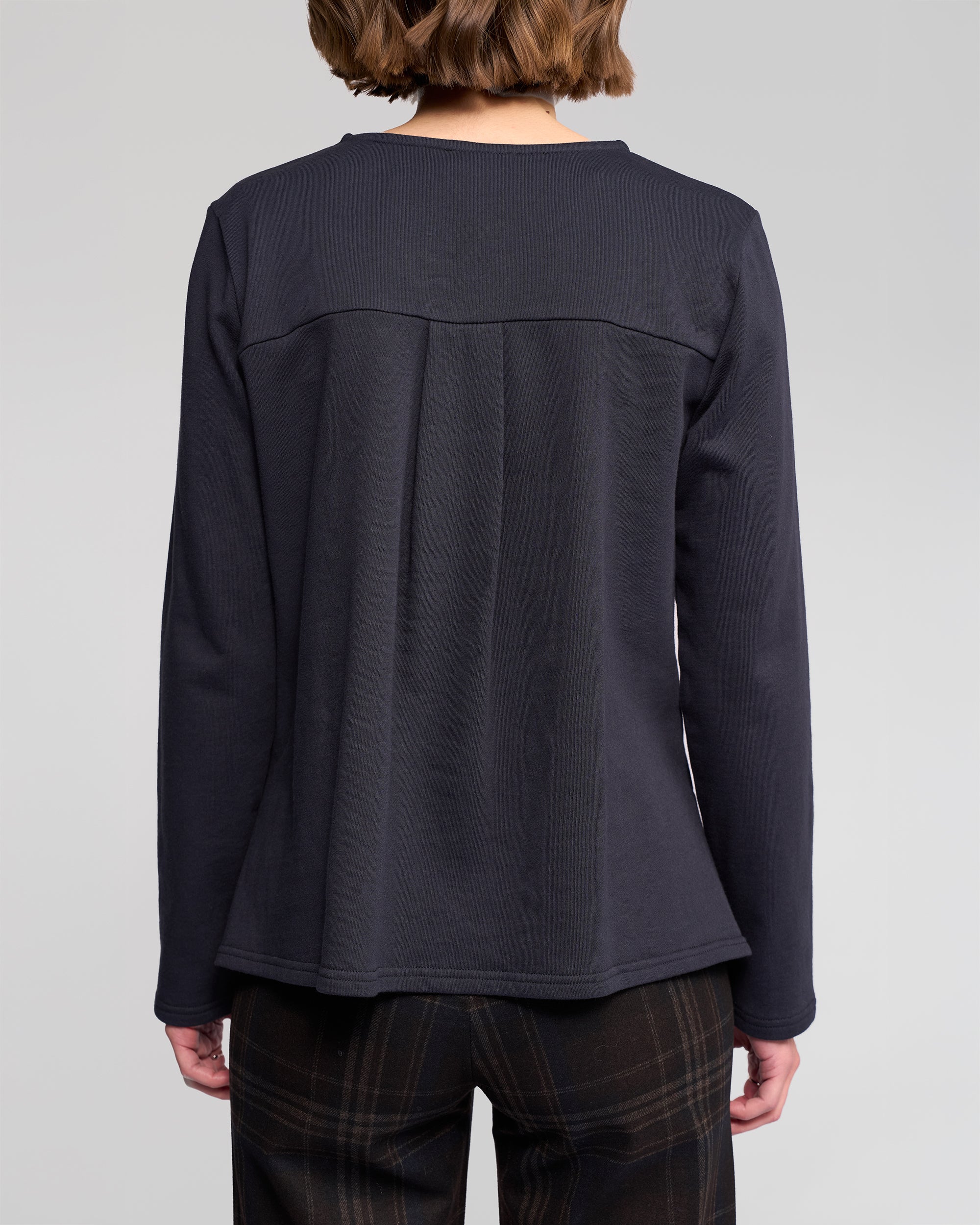A person with short brown hair, seen from behind, is wearing the Vassalli Chase Top in Ink and dark plaid winter-weight cotton pants against a plain light gray background.