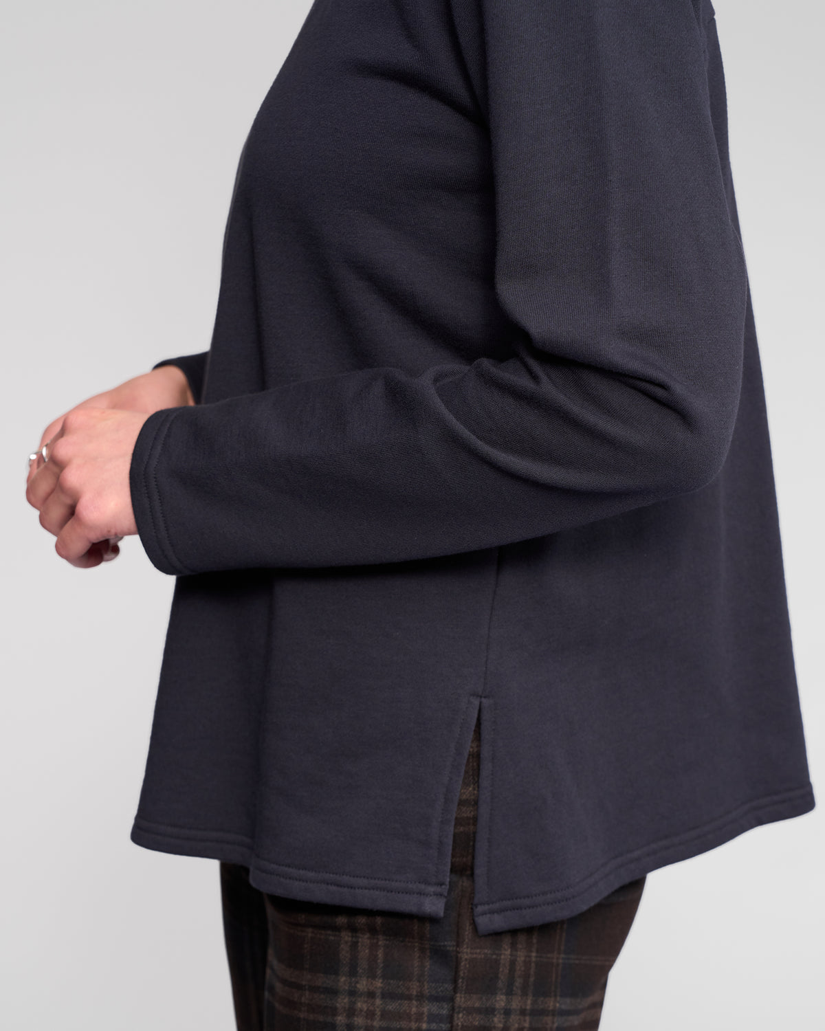 A person wears the Vassalli Chase Top - Ink, a relaxed-fit long sleeve navy sweatshirt with a side slit, paired with plaid pants. They stand sideways with hands loosely clasped against a plain light background.