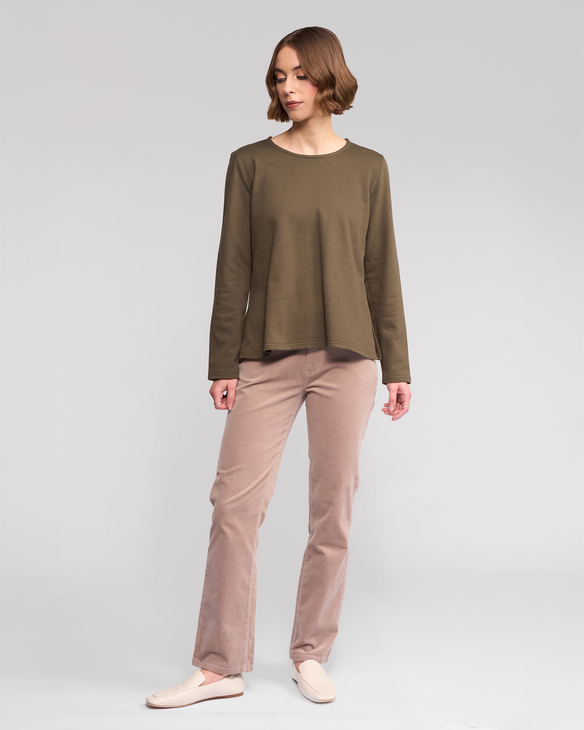 A woman with short brown hair wears the Vassalli Chase Top in Moss— a relaxed-fit, winter-weight cotton long sleeve—paired with light brown pants and cream slip-on shoes, standing against a plain light gray background.