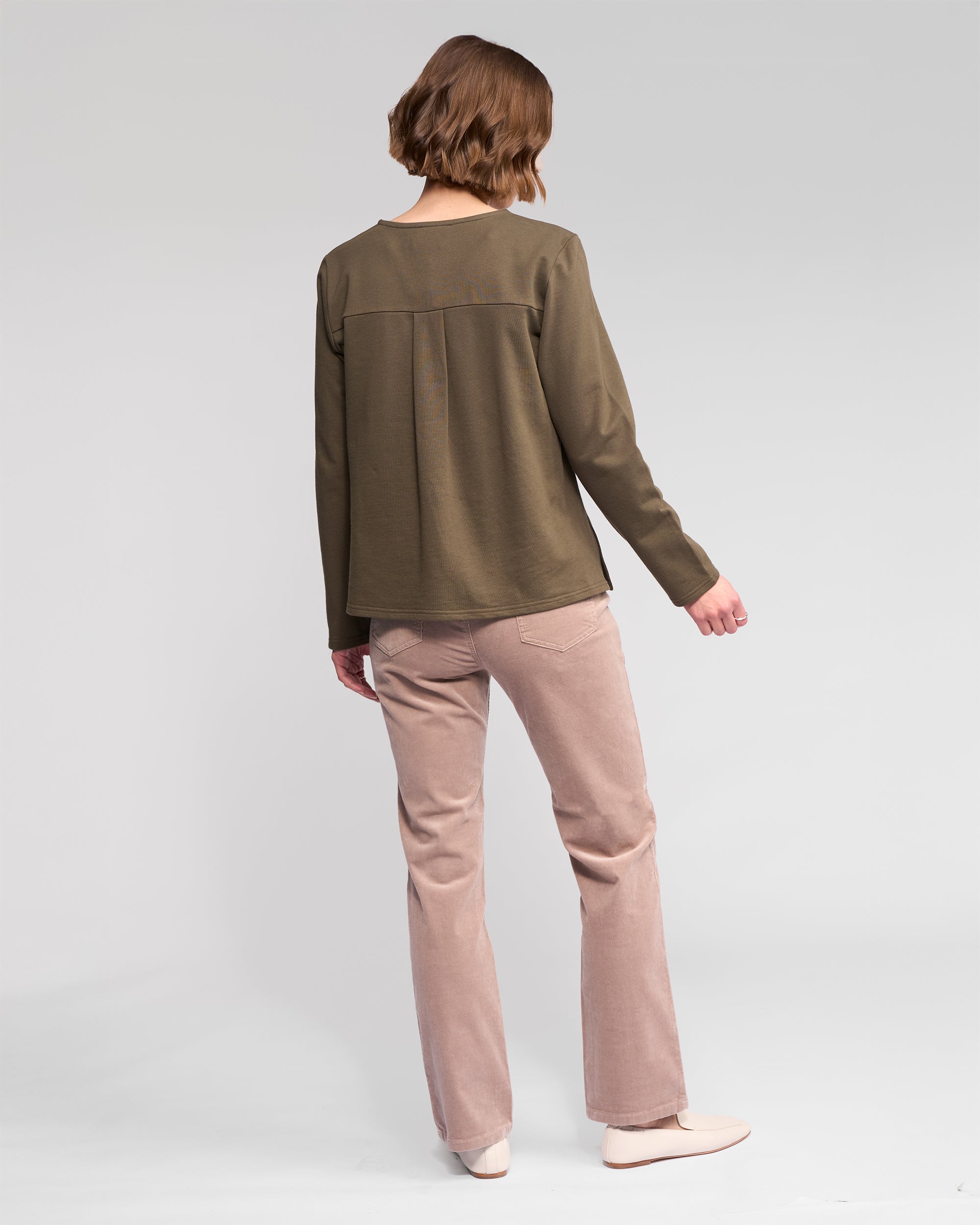 A person with short brown hair, facing away, wears the Vassalli Chase Top - Moss (a relaxed-fit winter-weight cotton long sleeve) with light pink pants and cream shoes against a plain light gray background.