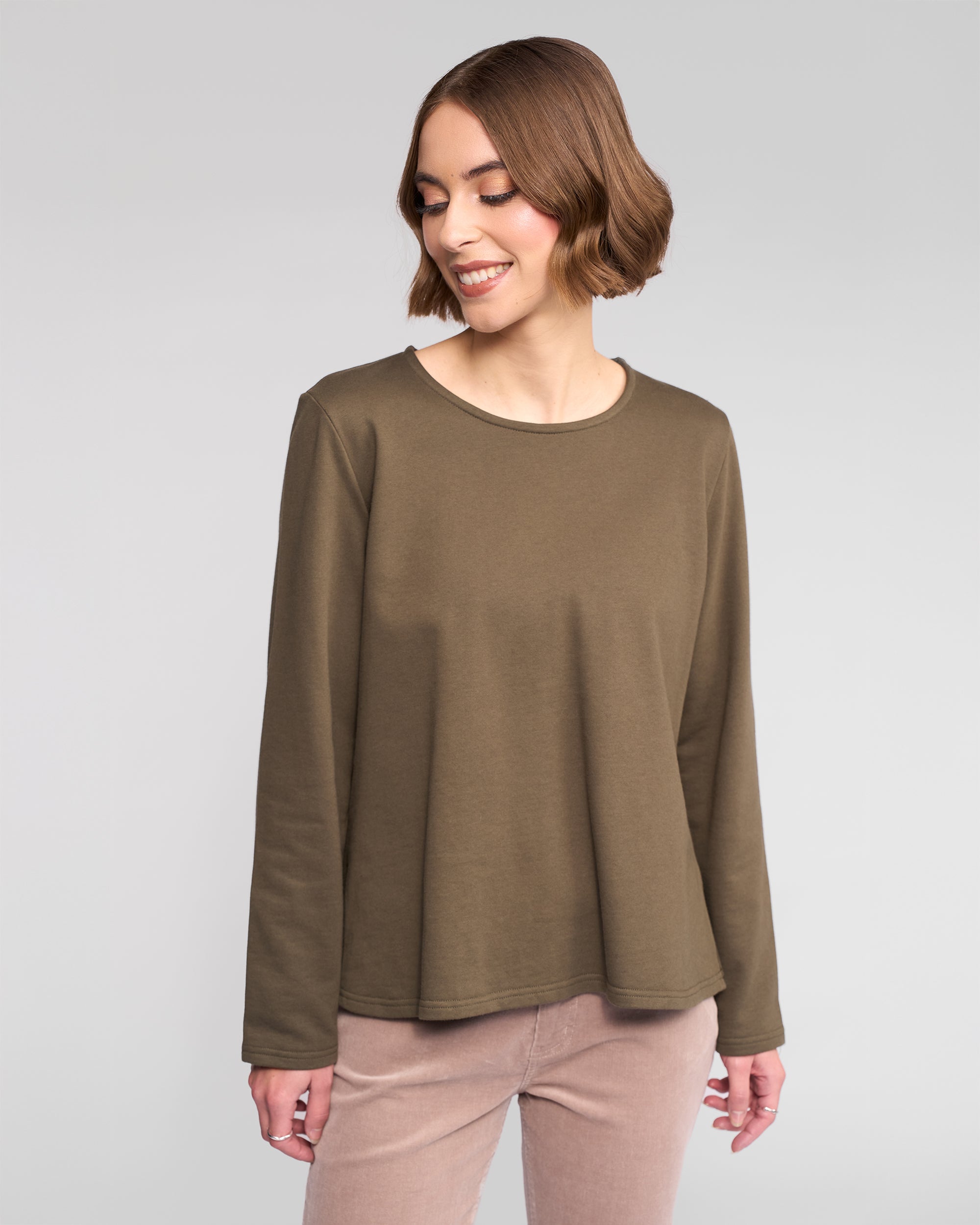 A woman with short brown hair smiles and looks down, wearing the Vassalli Chase Top in moss green and light beige pants against a plain light gray background.