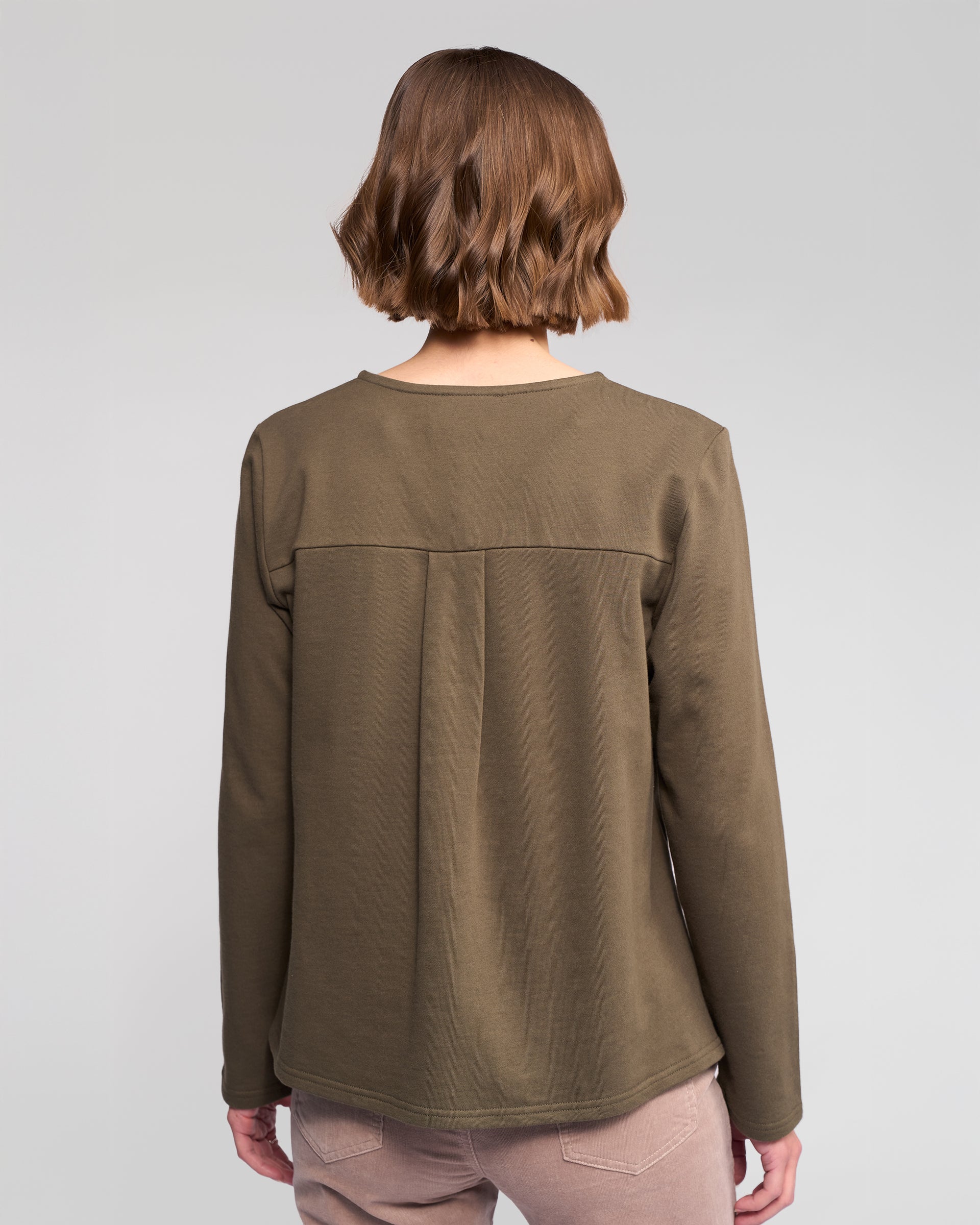 A person with short, wavy brown hair is seen from behind wearing the Vassalli Chase Top in Moss and light pink pants, standing against a plain light gray background.