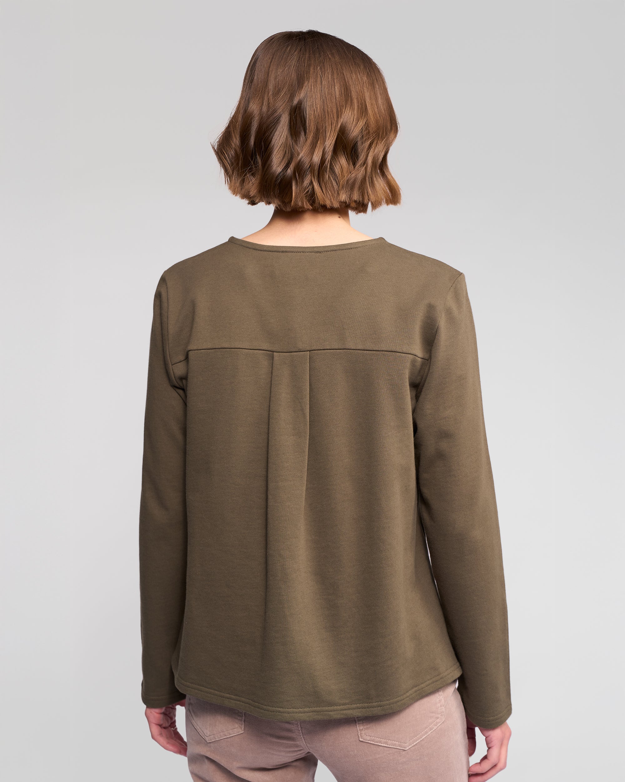 A person with short, wavy brown hair is seen from behind wearing the Vassalli Chase Top in Moss and light pink pants, standing against a plain light gray background.