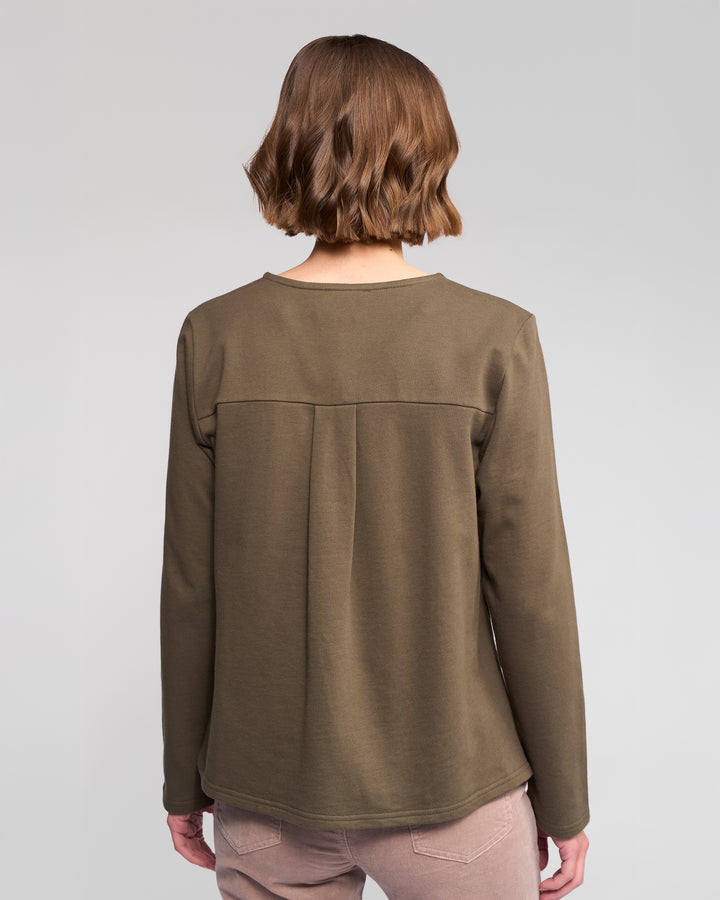 A person with short, wavy brown hair is seen from behind wearing the Vassalli Chase Top in Moss and light pink pants, standing against a plain light gray background.