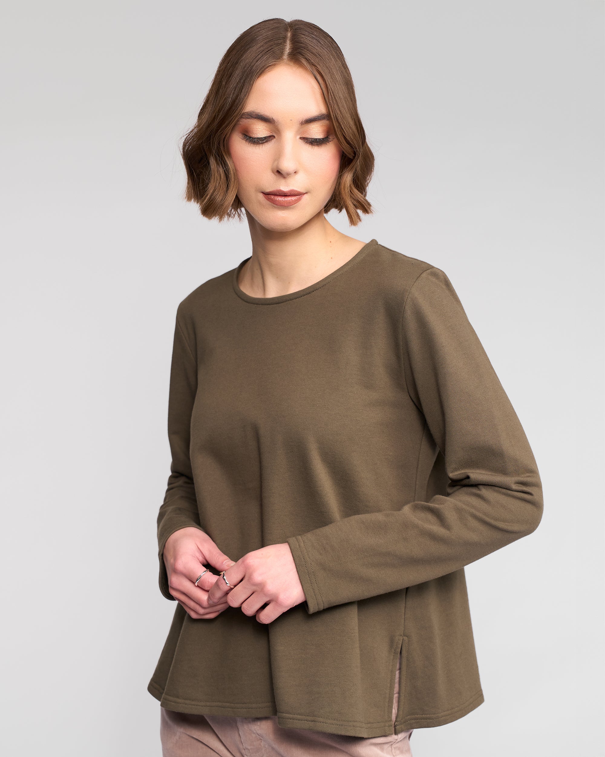 A woman with short brown hair wears the Vassalli Chase Top in Moss, a winter-weight cotton relaxed-fit long sleeve, paired with light beige pants. She looks down softly against a plain light gray background.