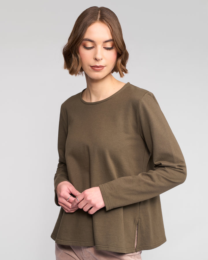 A woman with short brown hair wears the Vassalli Chase Top in Moss, a winter-weight cotton relaxed-fit long sleeve, paired with light beige pants. She looks down softly against a plain light gray background.