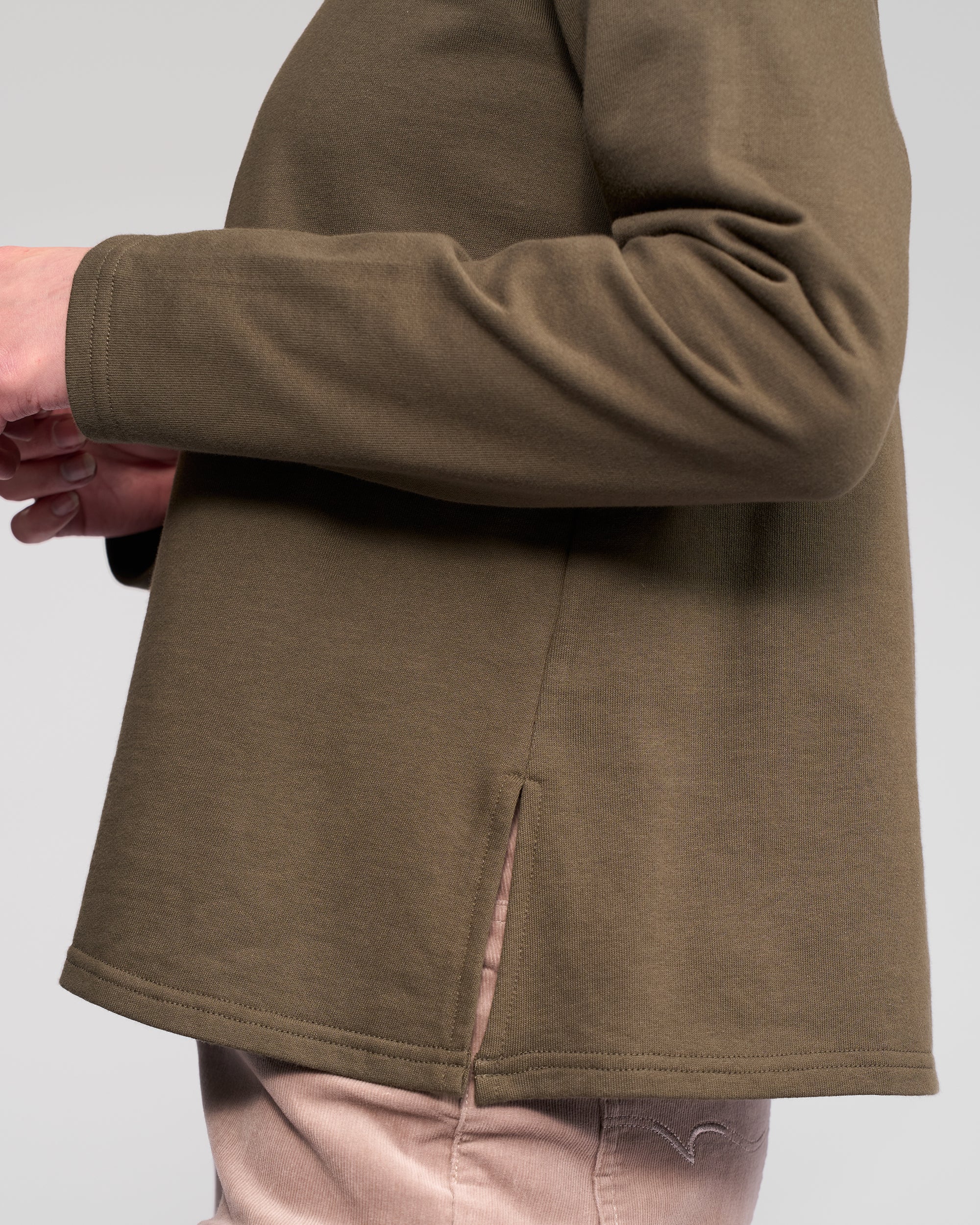 A person wears the Vassalli Chase Top in Moss, a relaxed-fit, winter-weight cotton long sleeve with a side slit at the hem, paired with light pants against a plain light gray background.