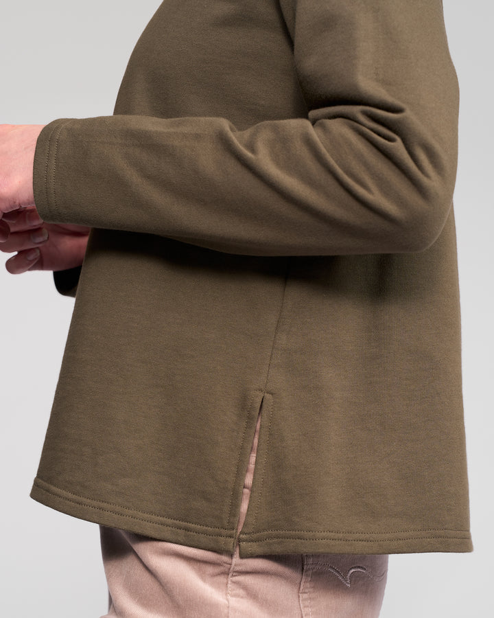 A person wears the Vassalli Chase Top in Moss, a relaxed-fit, winter-weight cotton long sleeve with a side slit at the hem, paired with light pants against a plain light gray background.