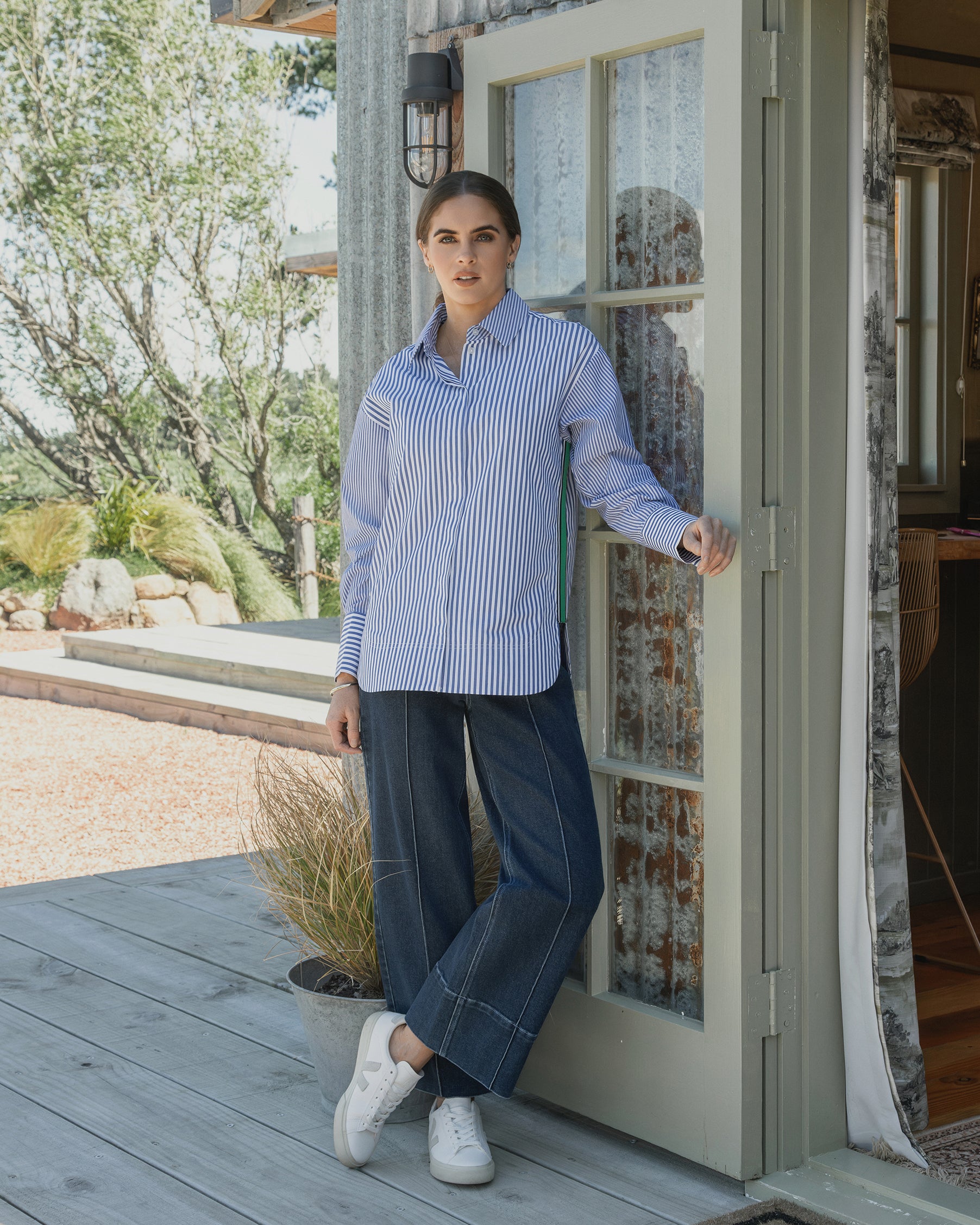 A woman stands on a wooden porch, leaning against a glass door. She wears the Vassalli Chart Shirt - Blue Stripe with wide-leg jeans and white sneakers. Trees and plants fill the background.