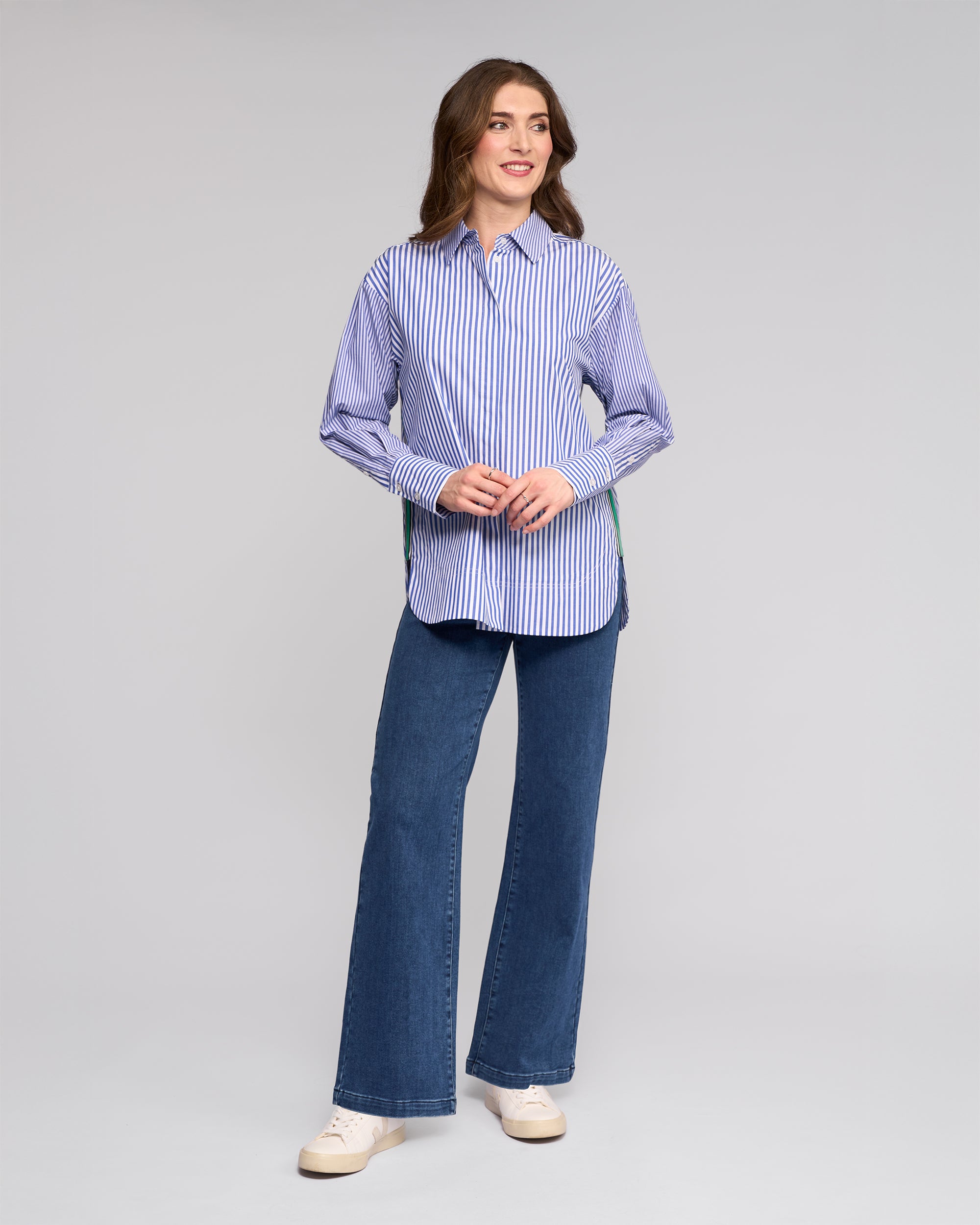 A woman smiles wearing the Vassalli Chart Shirt in Blue Stripe, an oversized cotton shirt, paired with blue wide-leg jeans and white sneakers against a plain light gray background.