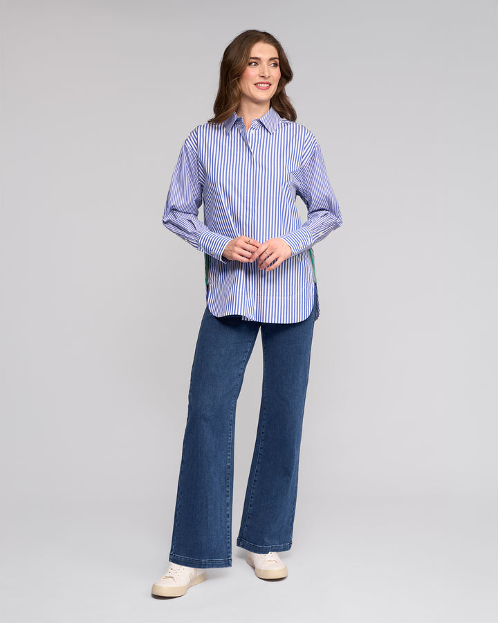 A woman smiles wearing the Vassalli Chart Shirt in Blue Stripe, an oversized cotton shirt, paired with blue wide-leg jeans and white sneakers against a plain light gray background.