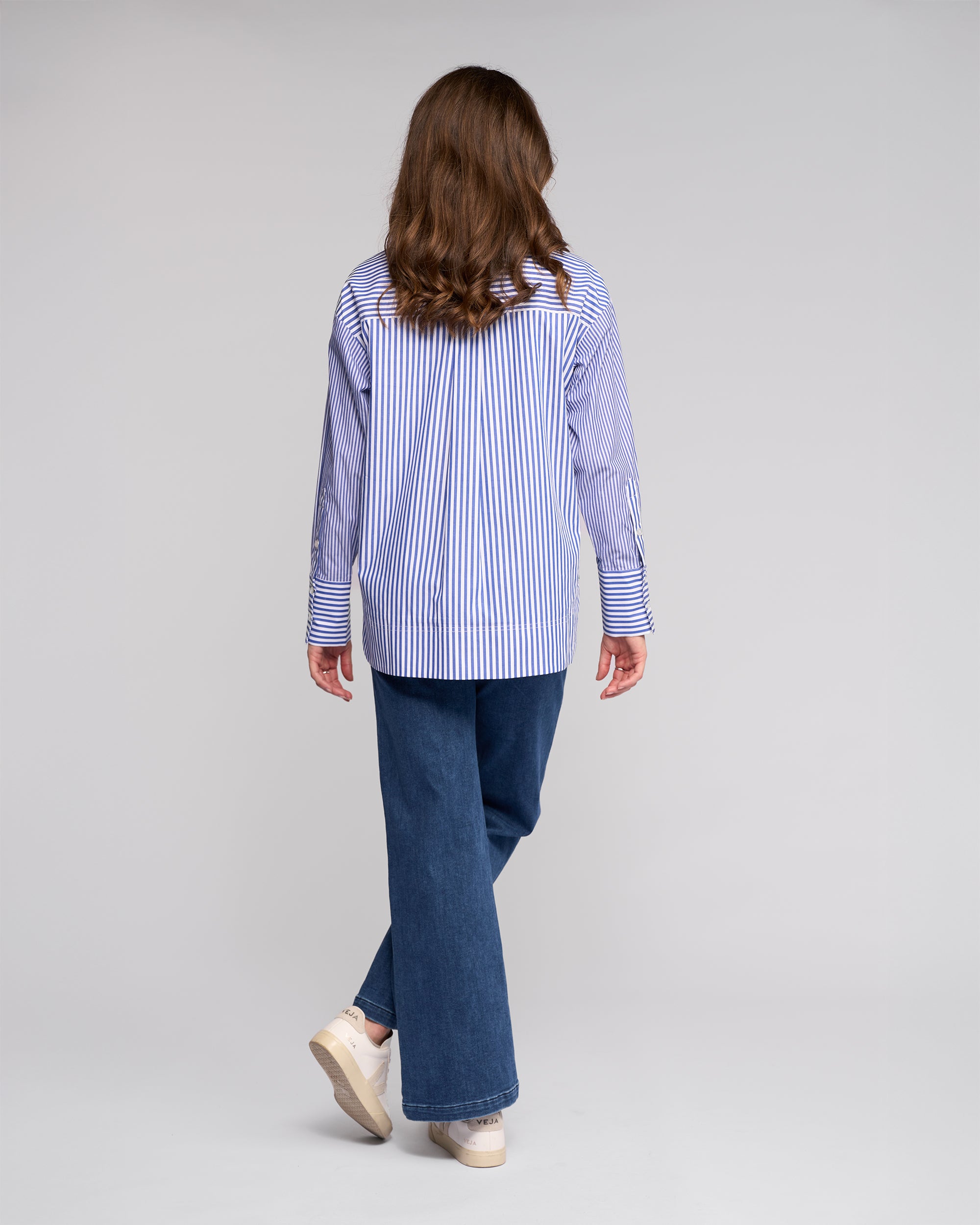 A person with long brown hair, wearing the Vassalli Chart Shirt - Blue Stripe, wide-leg blue jeans, and beige platform shoes, stands facing away from the camera against a plain light background.