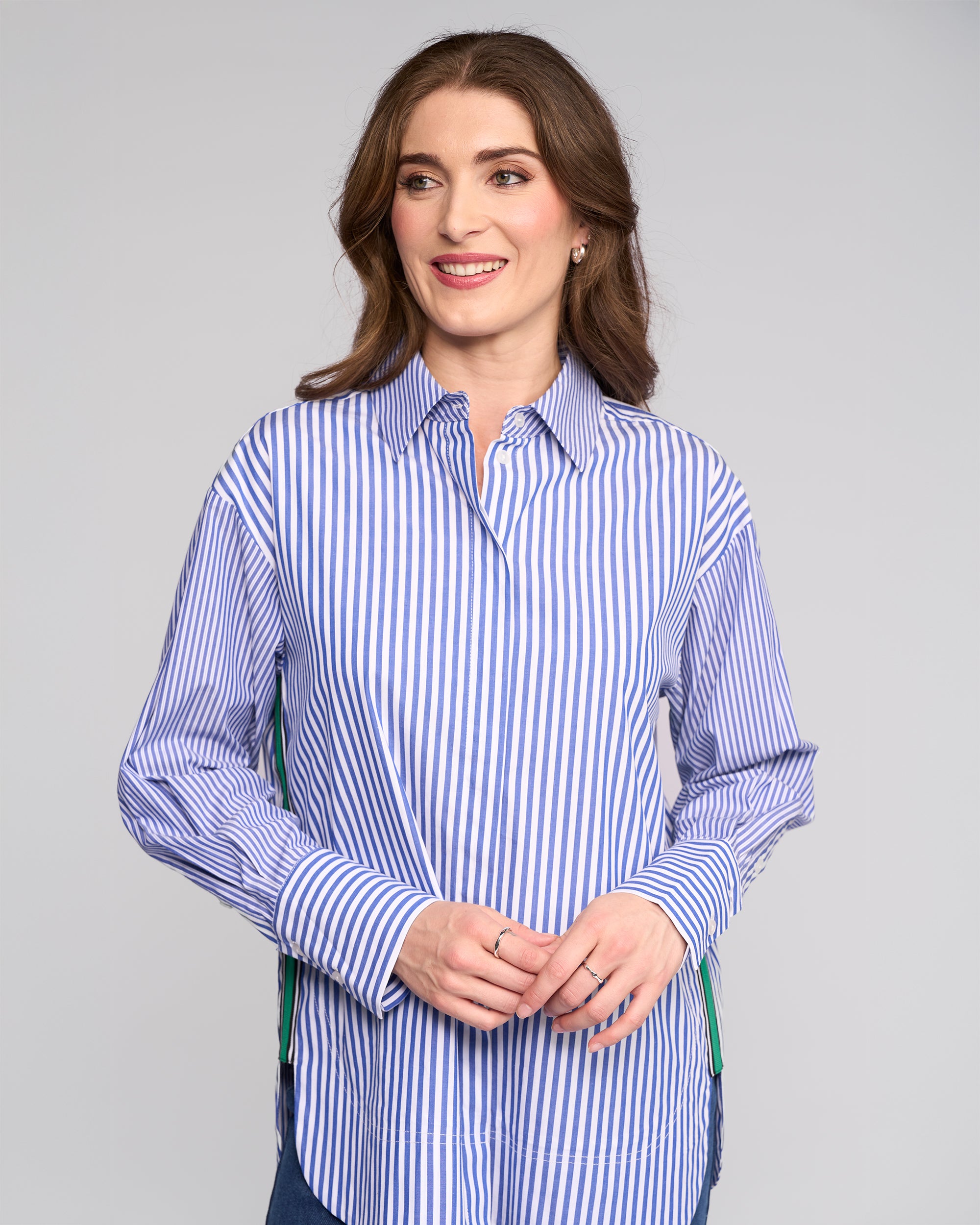 A woman with brown hair smiles gently in the Vassalli Chart Shirt - Blue Stripe, an oversized cotton piece, standing against a plain light gray background with her hands loosely clasped.