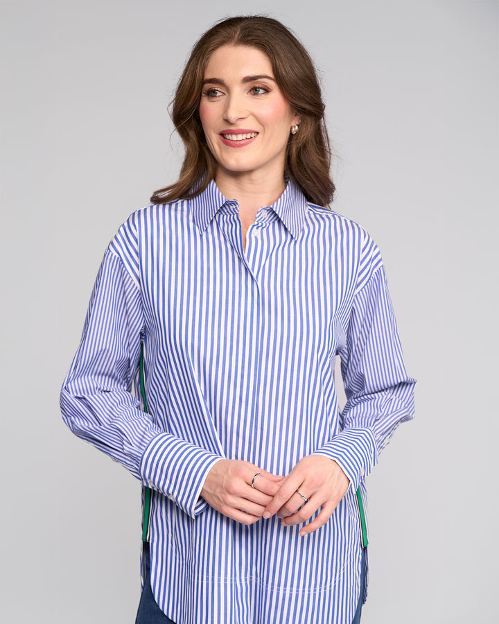 A woman with brown hair smiles gently in the Vassalli Chart Shirt - Blue Stripe, an oversized cotton piece, standing against a plain light gray background with her hands loosely clasped.