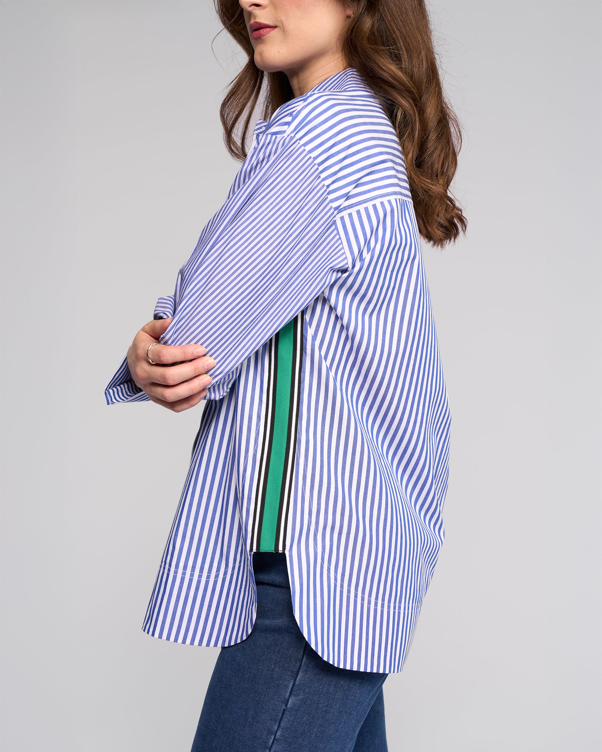 Model wears the Vassalli Chart Shirt - Blue Stripe, an oversized cotton piece with side green and black stripes, paired with blue jeans. Posed against a plain gray backdrop, only the upper body is shown for a modern shirting look.