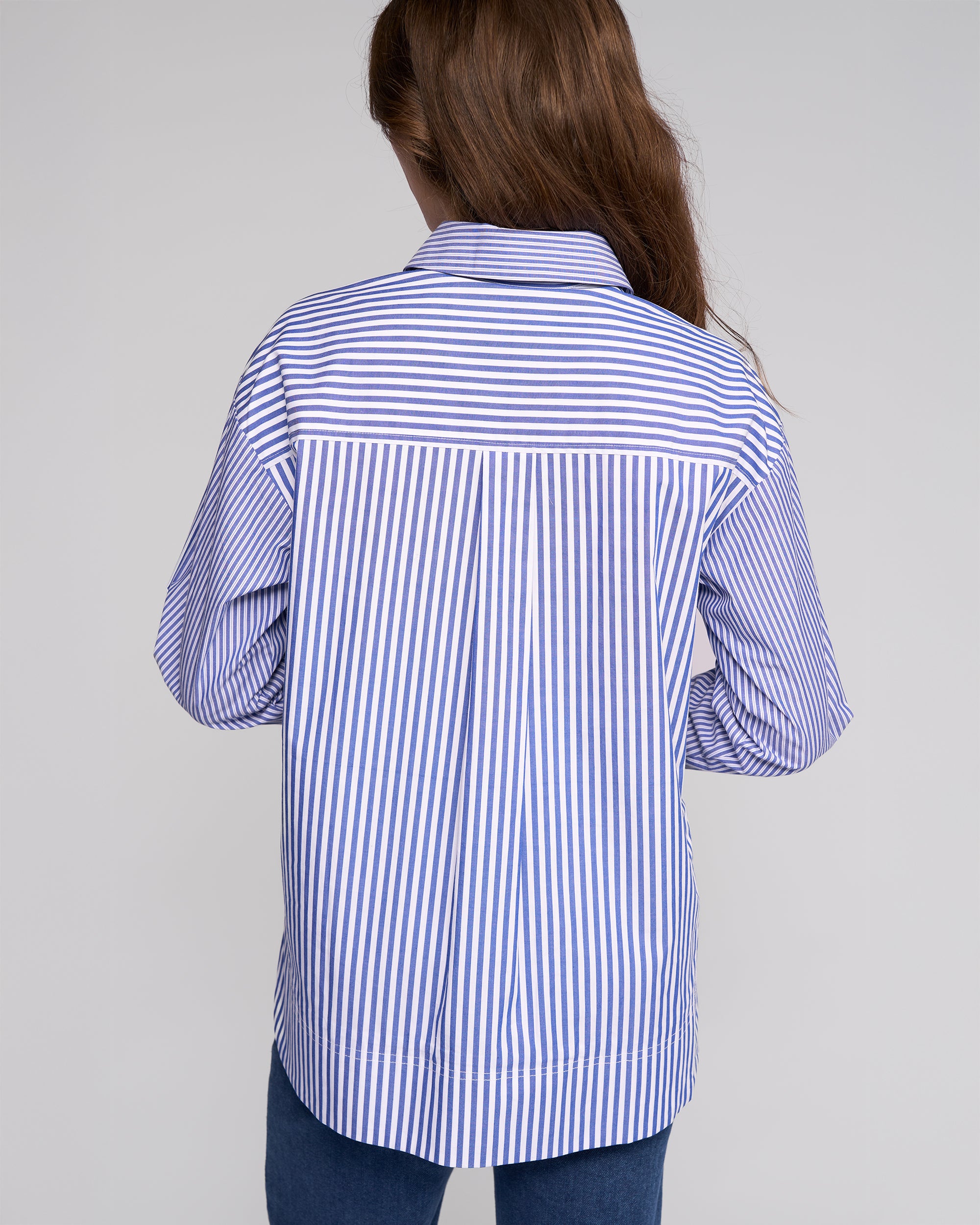 A person with long brown hair, seen from behind against a light gray background, models the Vassalli Chart Shirt - Blue Stripe, an oversized cotton striped shirt paired with dark blue jeans for a modern shirting look.