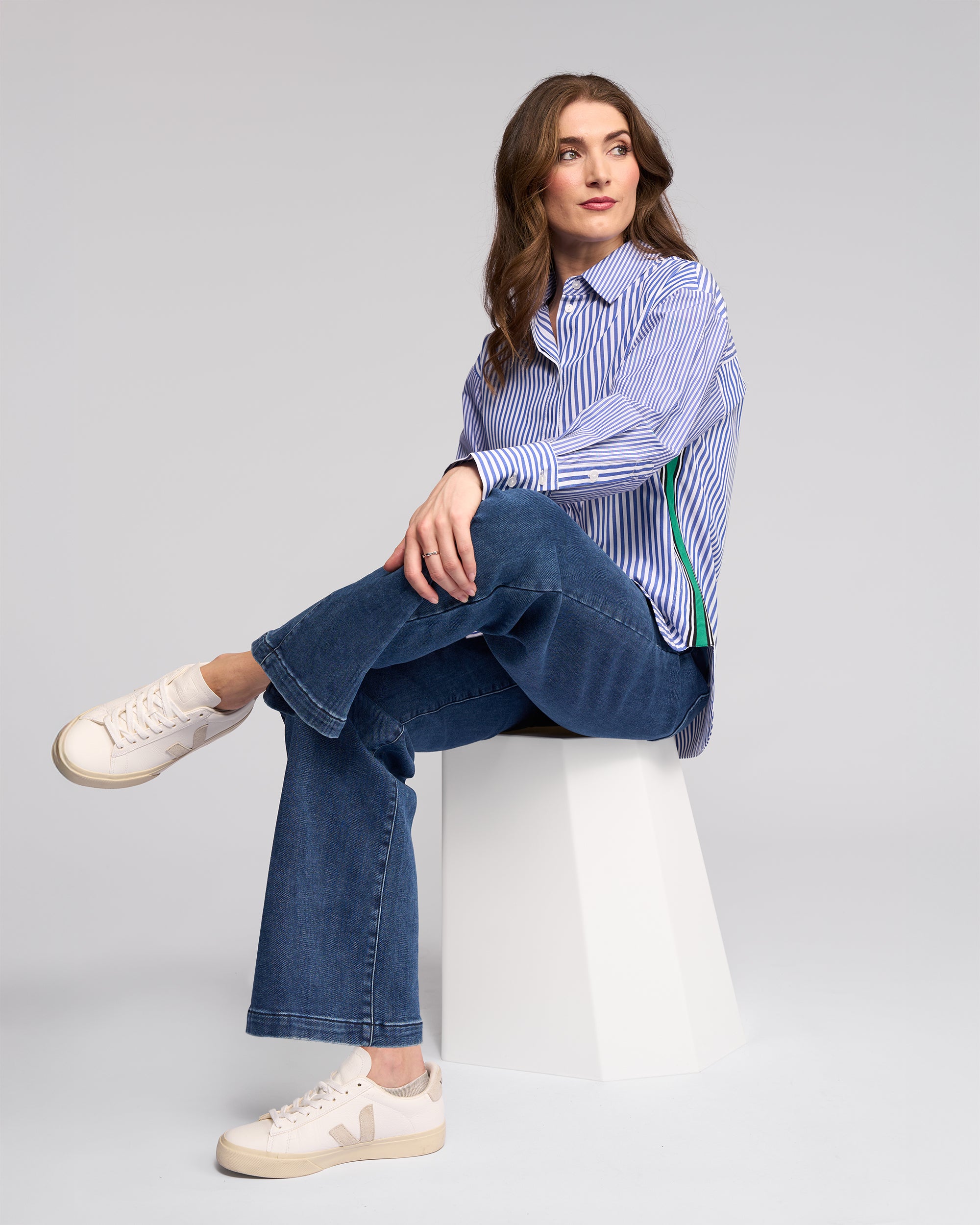 A woman with long brown hair sits on a white geometric stool, wearing the Vassalli Chart Shirt - Blue Stripe, blue jeans, and white sneakers. She gazes to the side with a relaxed expression against a plain background.