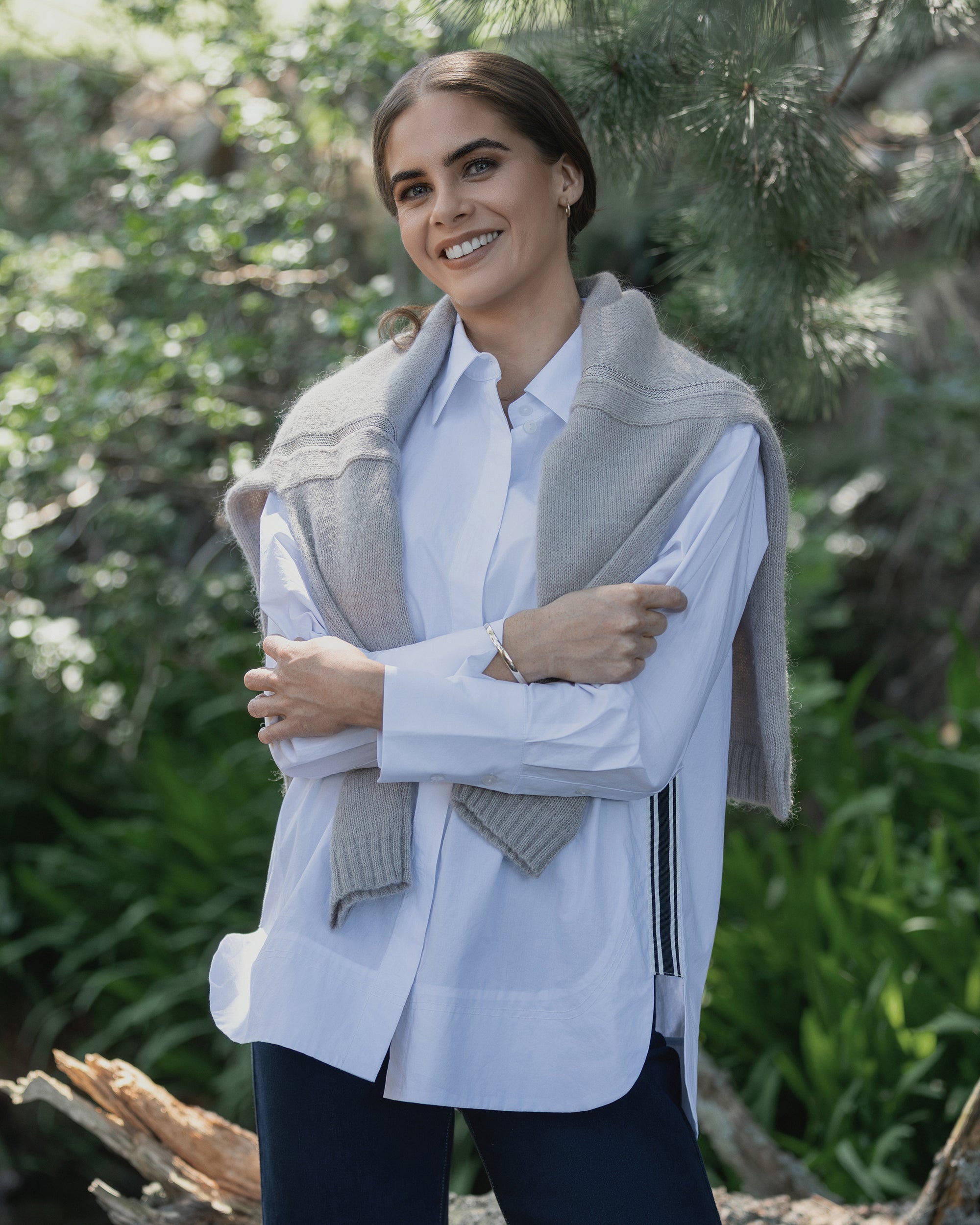 A woman with brown hair smiles outdoors, arms crossed, wearing the Vassalli Chart Shirt in white and a gray sweater over her shoulders, surrounded by greenery and sunlight.