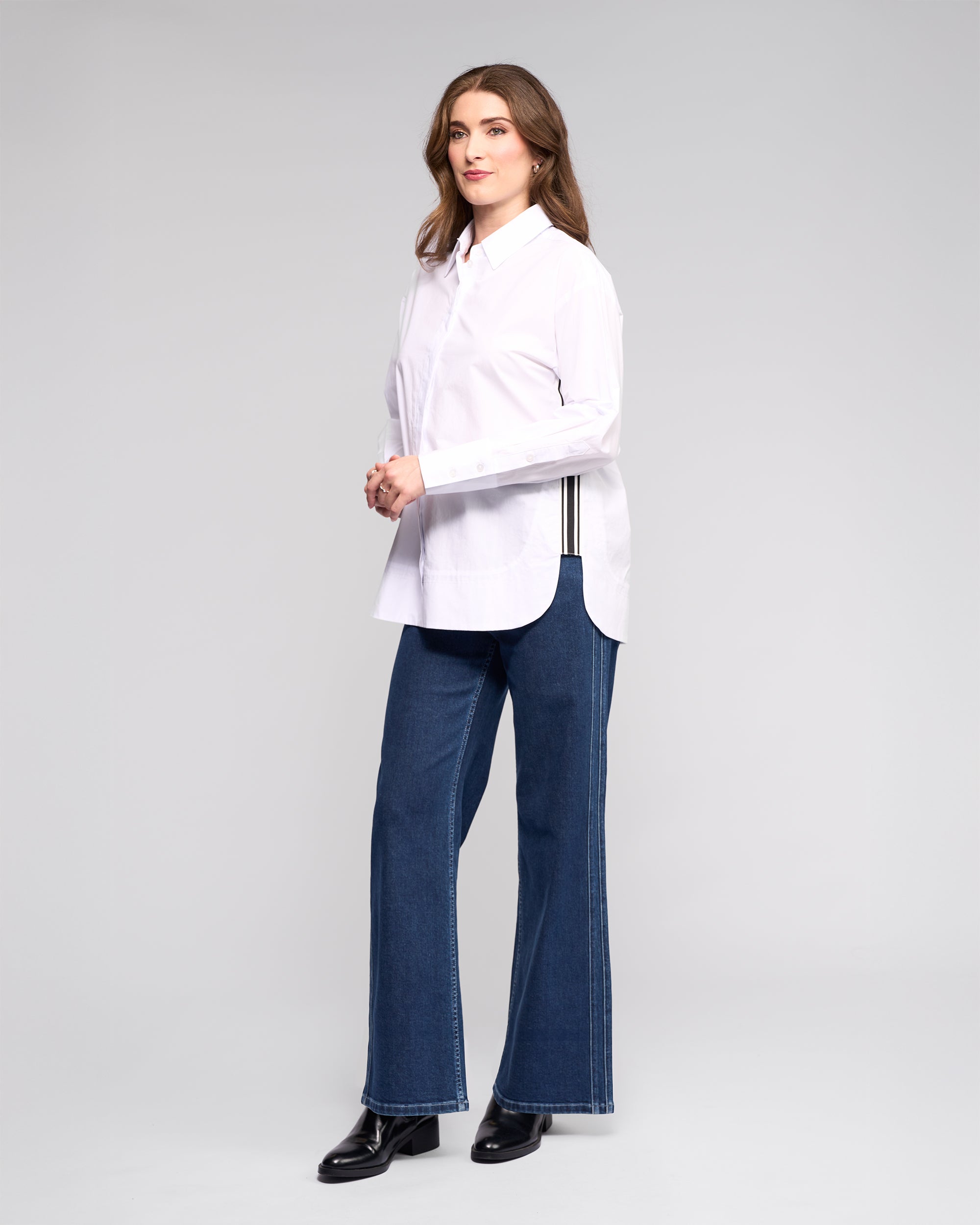 A woman models the Vassalli Chart Shirt in white, an oversized cotton top with black side stripes, paired with blue jeans and black boots. Her wavy brown hair and neutral gaze embody modern shirting style in a studio setting.