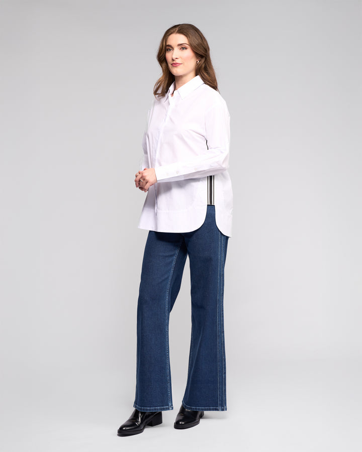 A woman models the Vassalli Chart Shirt in white, an oversized cotton top with black side stripes, paired with blue jeans and black boots. Her wavy brown hair and neutral gaze embody modern shirting style in a studio setting.