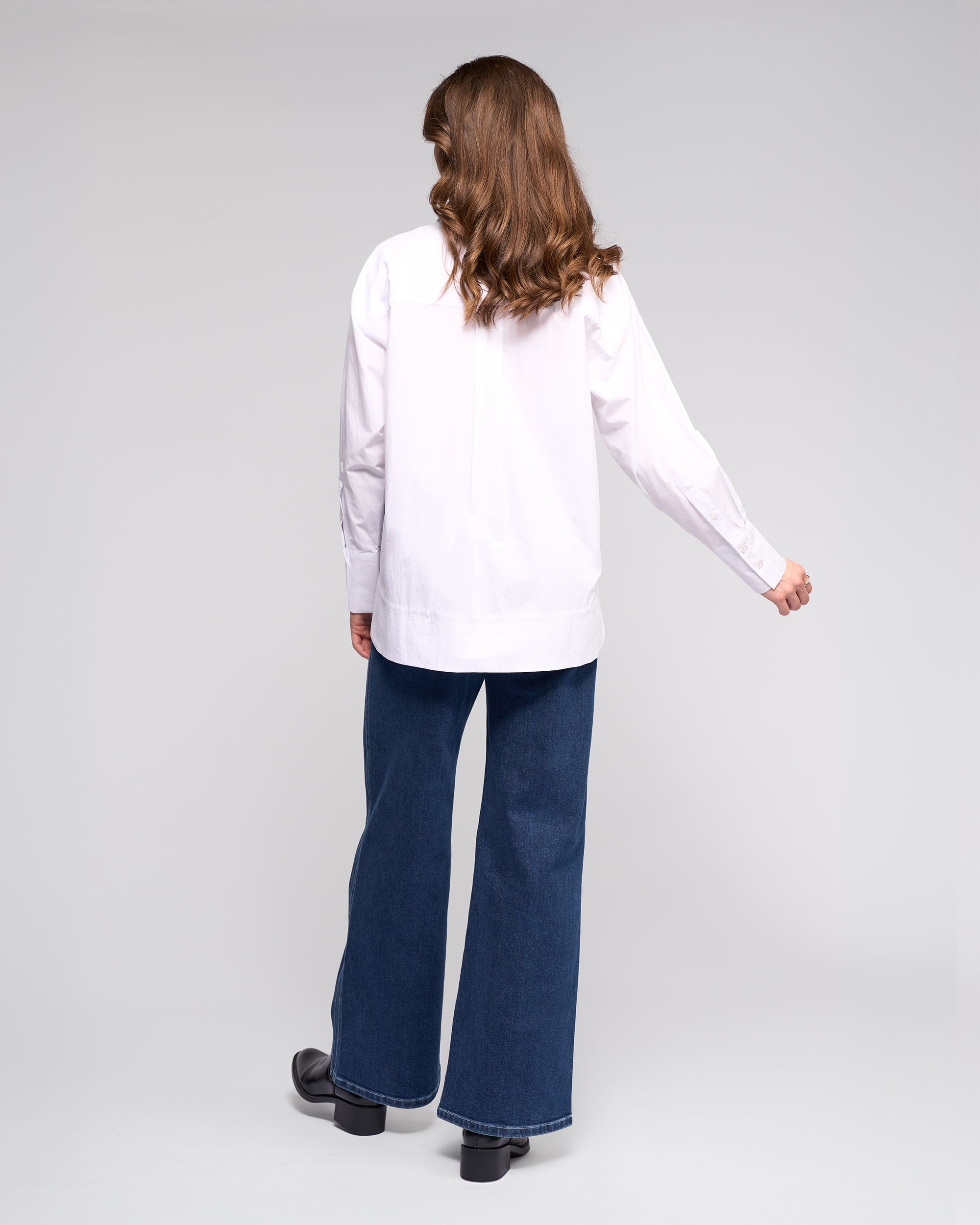 A person with wavy brown hair faces away, wearing the Vassalli Chart Shirt in white, paired with wide-leg blue jeans and black shoes, against a plain light background.