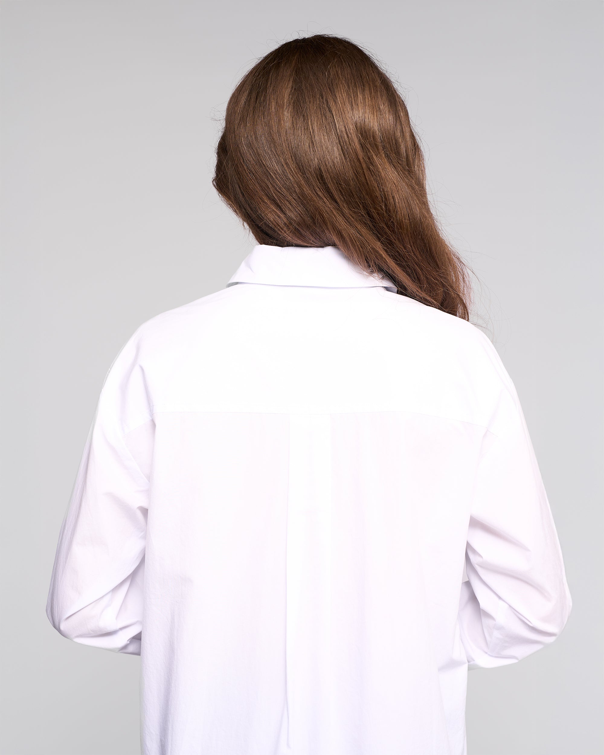 A person with long brown hair, wearing the Vassalli Chart Shirt - White, stands with their back to the camera against a plain light background.