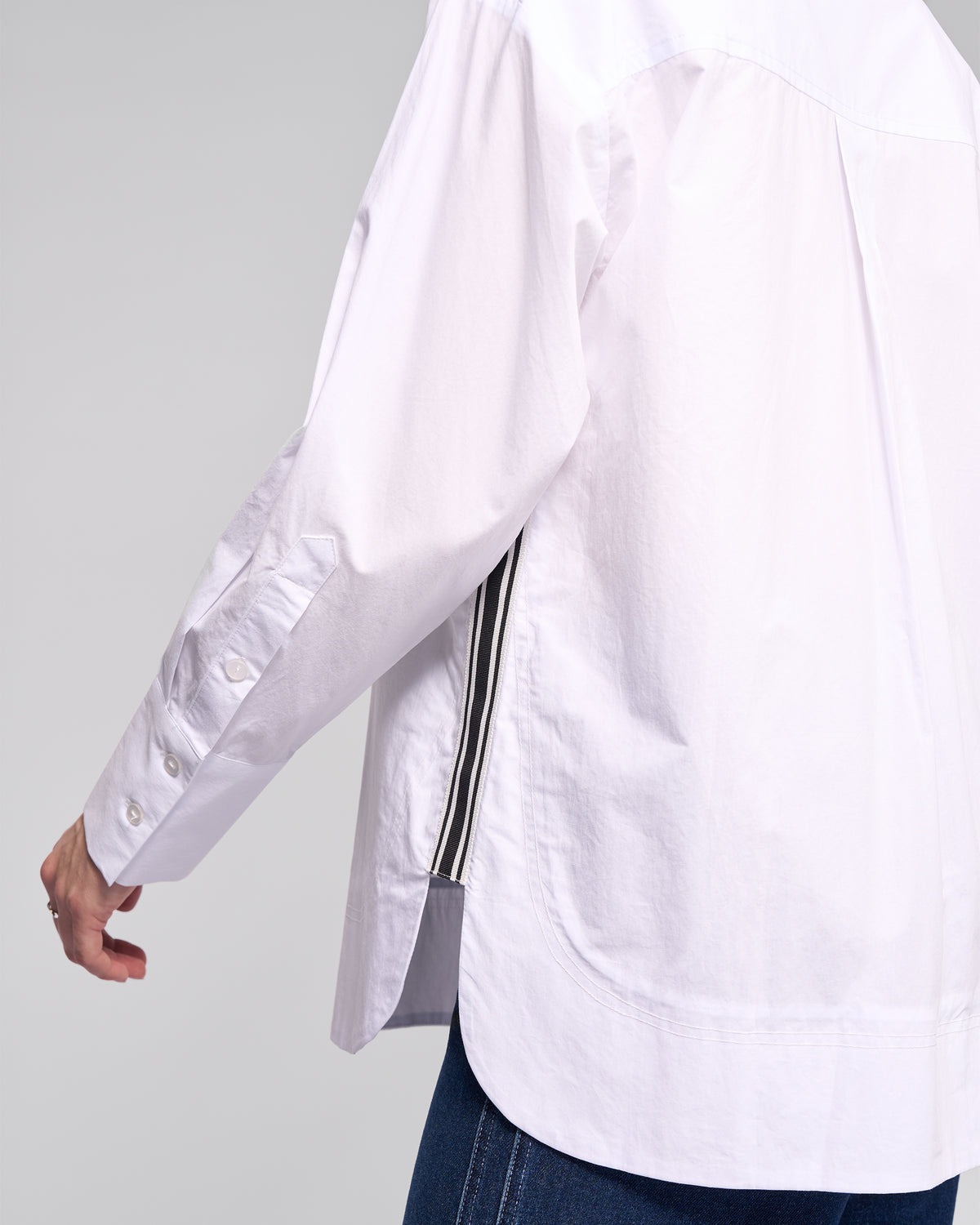 A person is shown from the back, wearing the Vassalli Chart Shirt in white—an oversized cotton style with black-and-white side stripes—paired with blue jeans, one arm slightly extended.