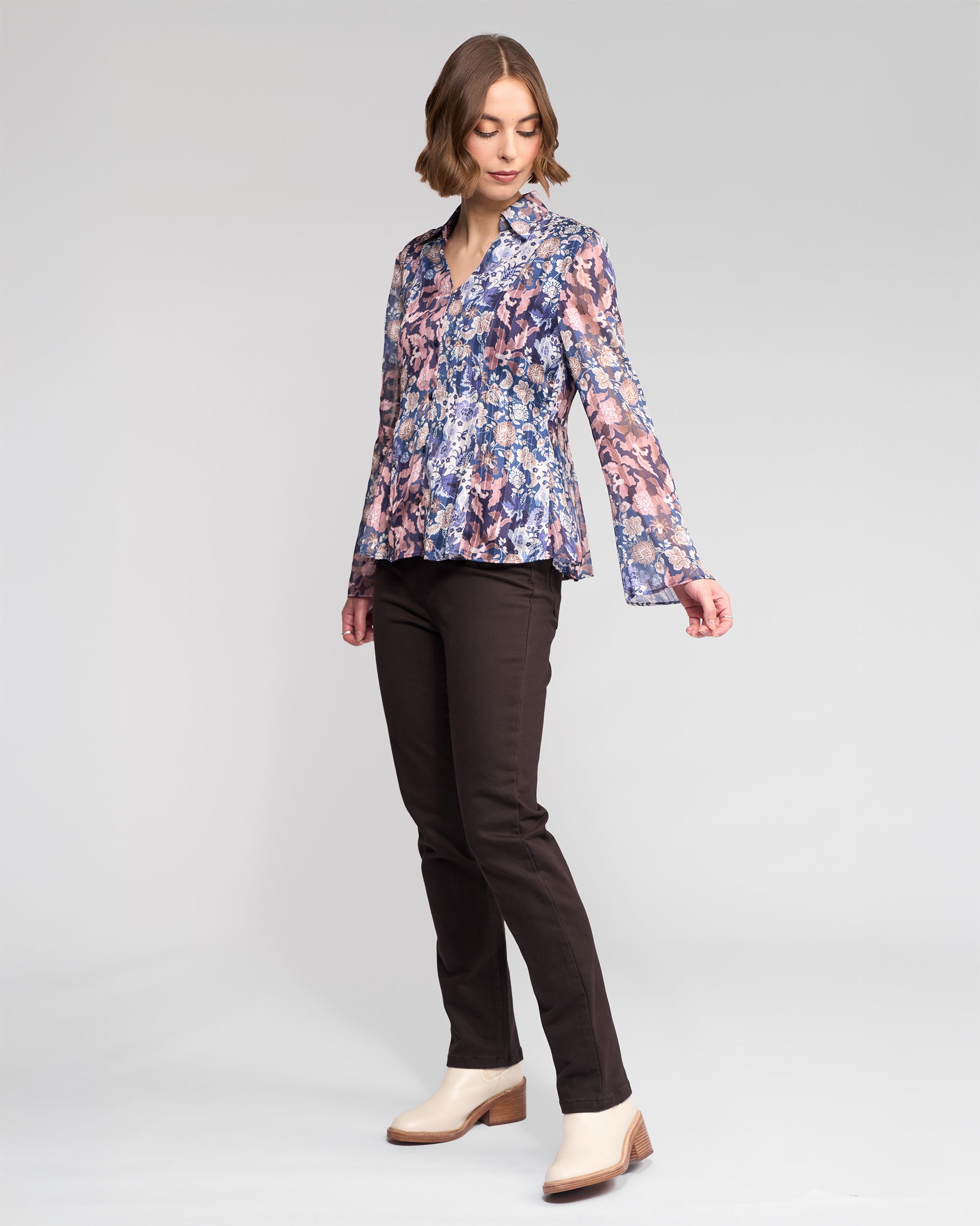 A woman poses in the Vassalli Valley Shirt - Rhea, a vibrant chiffon, long-sleeve top with a multicolored floral design, styled with dark brown pants and light shoes against a plain light gray background.