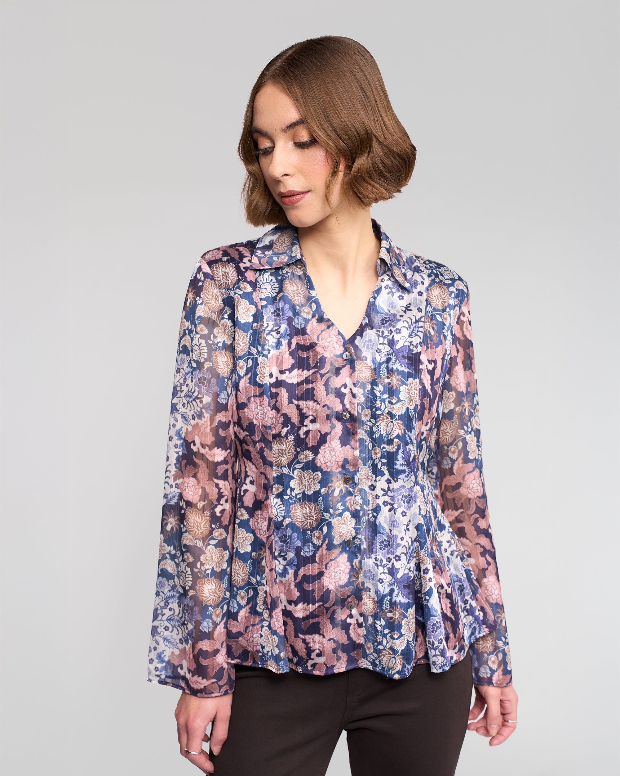 A woman with short brown hair wears the Vassalli Valley Shirt - Rhea, a long-sleeve blouse with blue, purple, and pink floral patterns. She looks down and slightly to the side against a plain light gray background.
