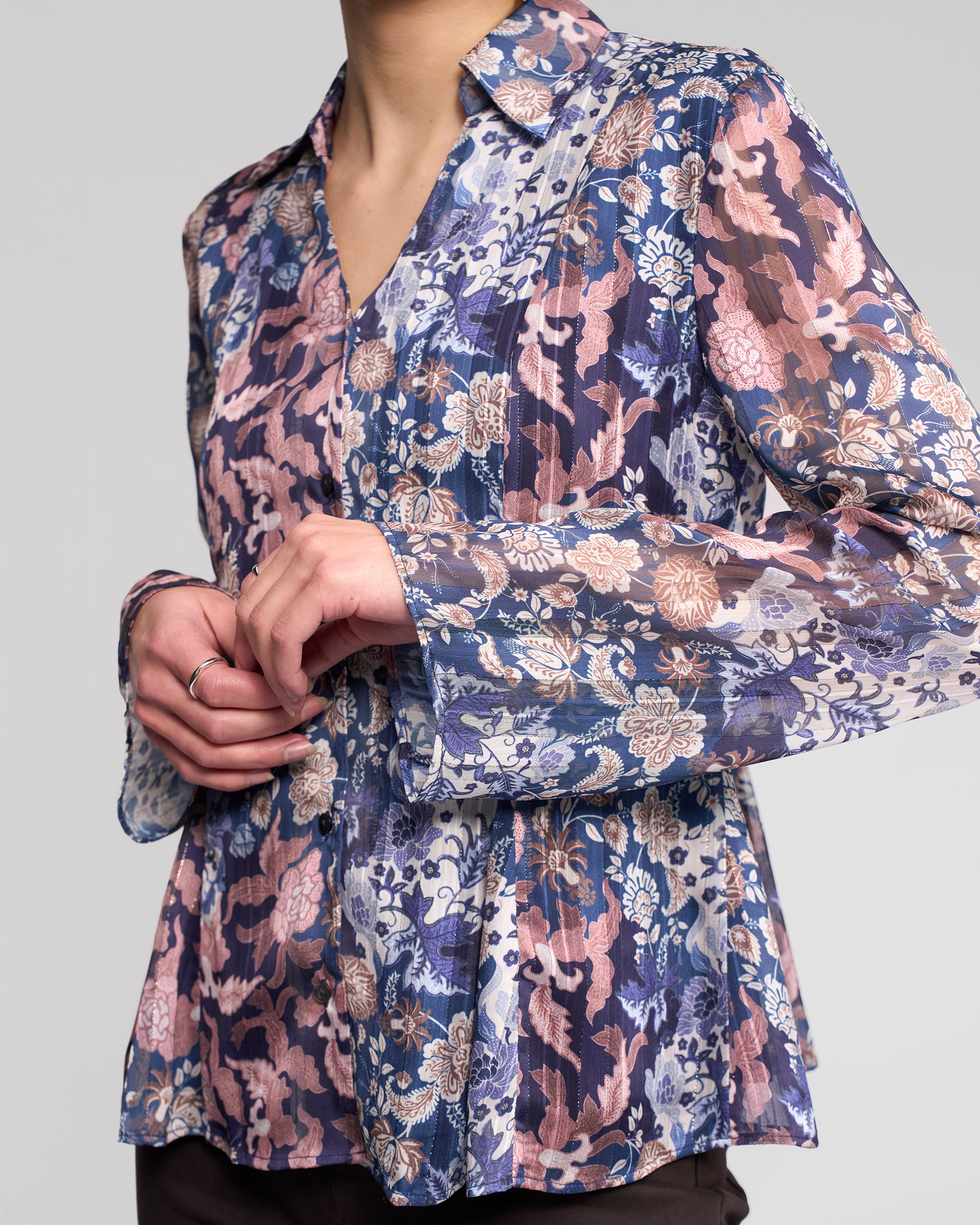 A person wearing the Vassalli Valley Shirt - Rhea, a long-sleeve floral blouse in blue, pink, and beige, stands with hands gently clasped. The face is not visible.