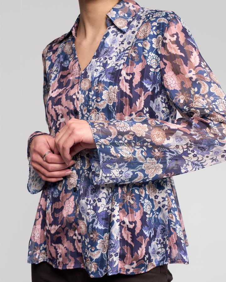 A person wearing the Vassalli Valley Shirt - Rhea, a long-sleeve floral blouse in blue, pink, and beige, stands with hands gently clasped. The face is not visible.