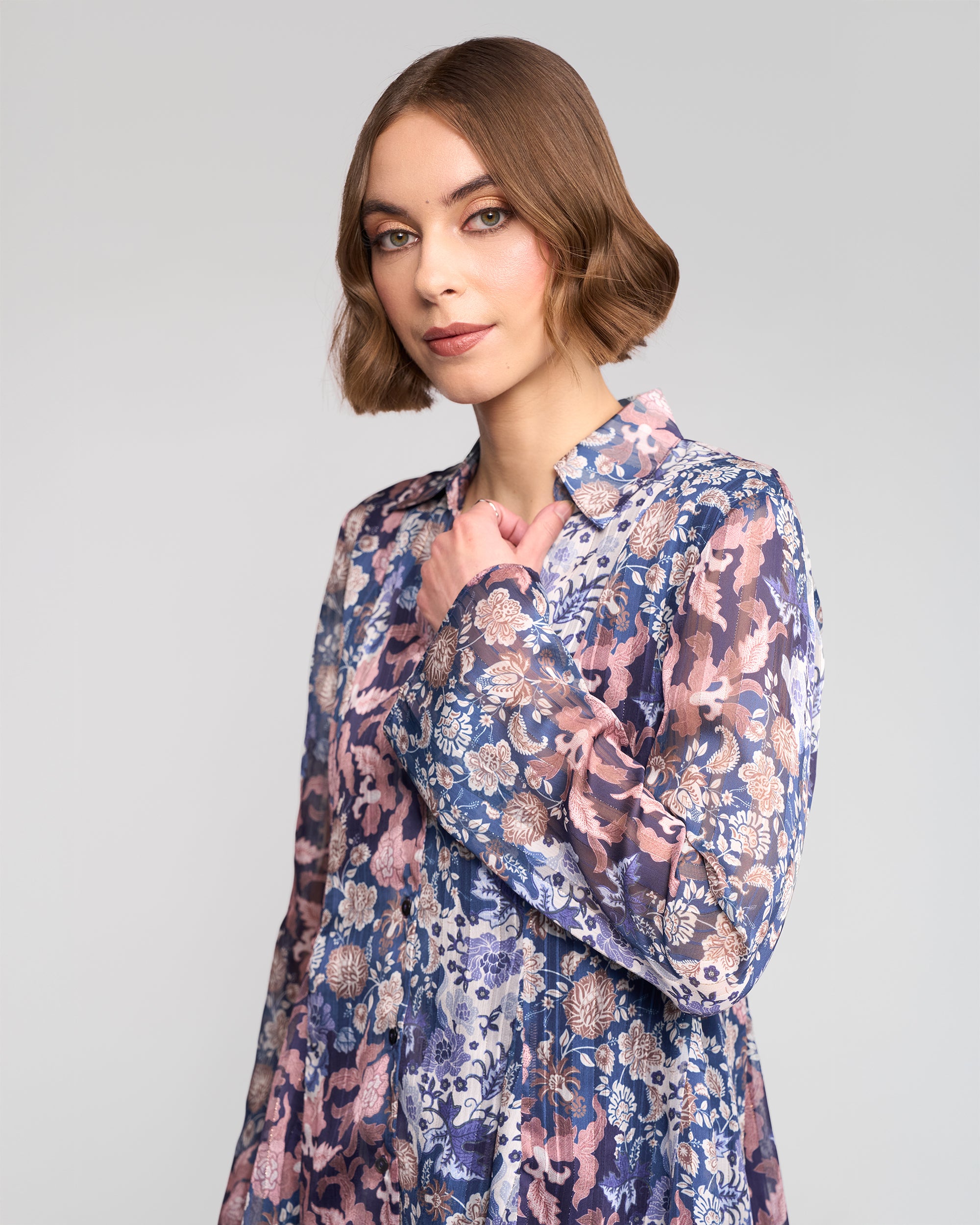A woman with short brown hair confidently models the Vassalli Valley Shirt - Rhea, a long-sleeve floral blouse, as she stands against a light gray background and looks directly at the camera.