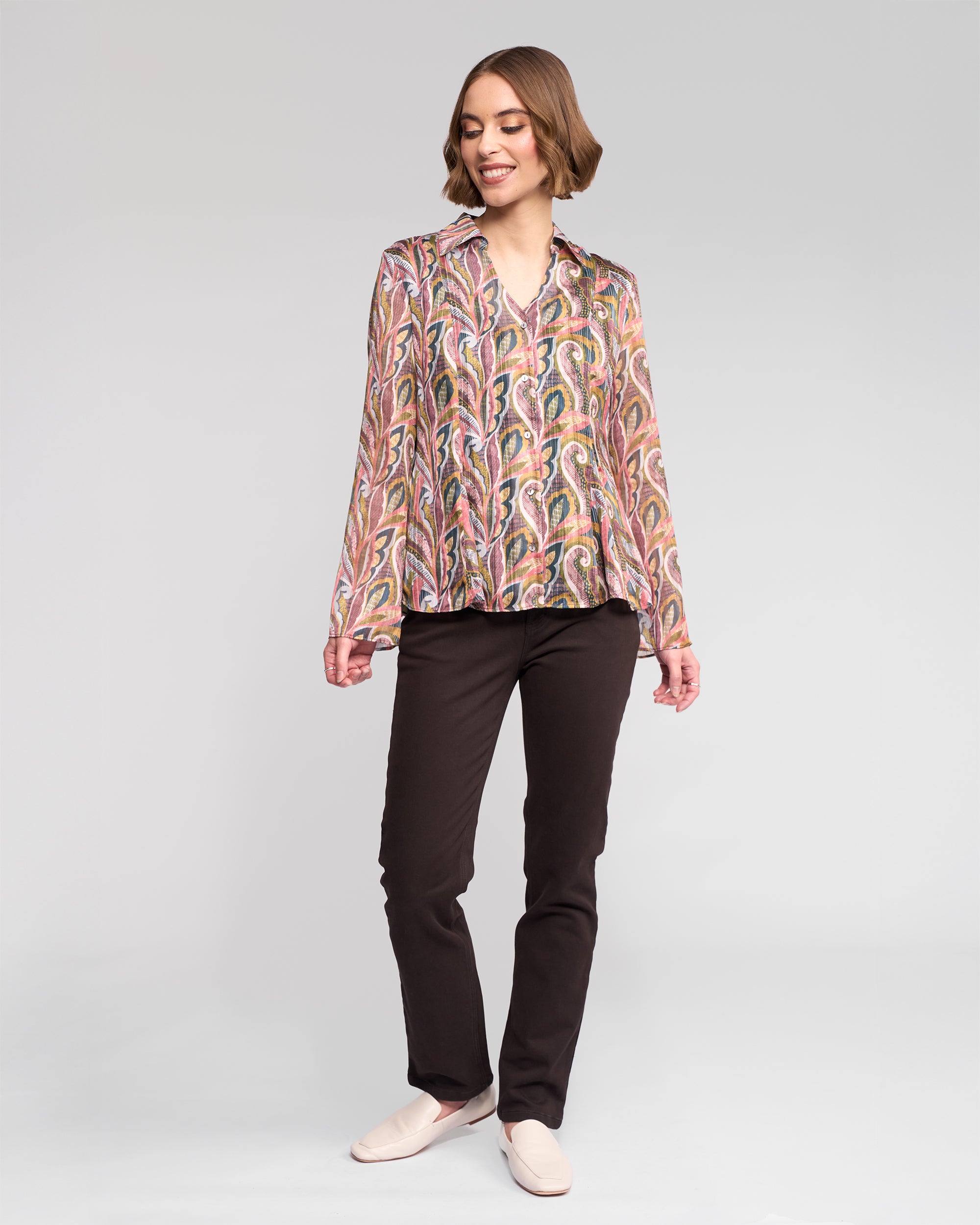 A woman with short hair, slightly turned to the side, smiles against a plain background wearing the Vassalli Valley Shirt in Sable, paired with dark pants and light slip-on shoes.