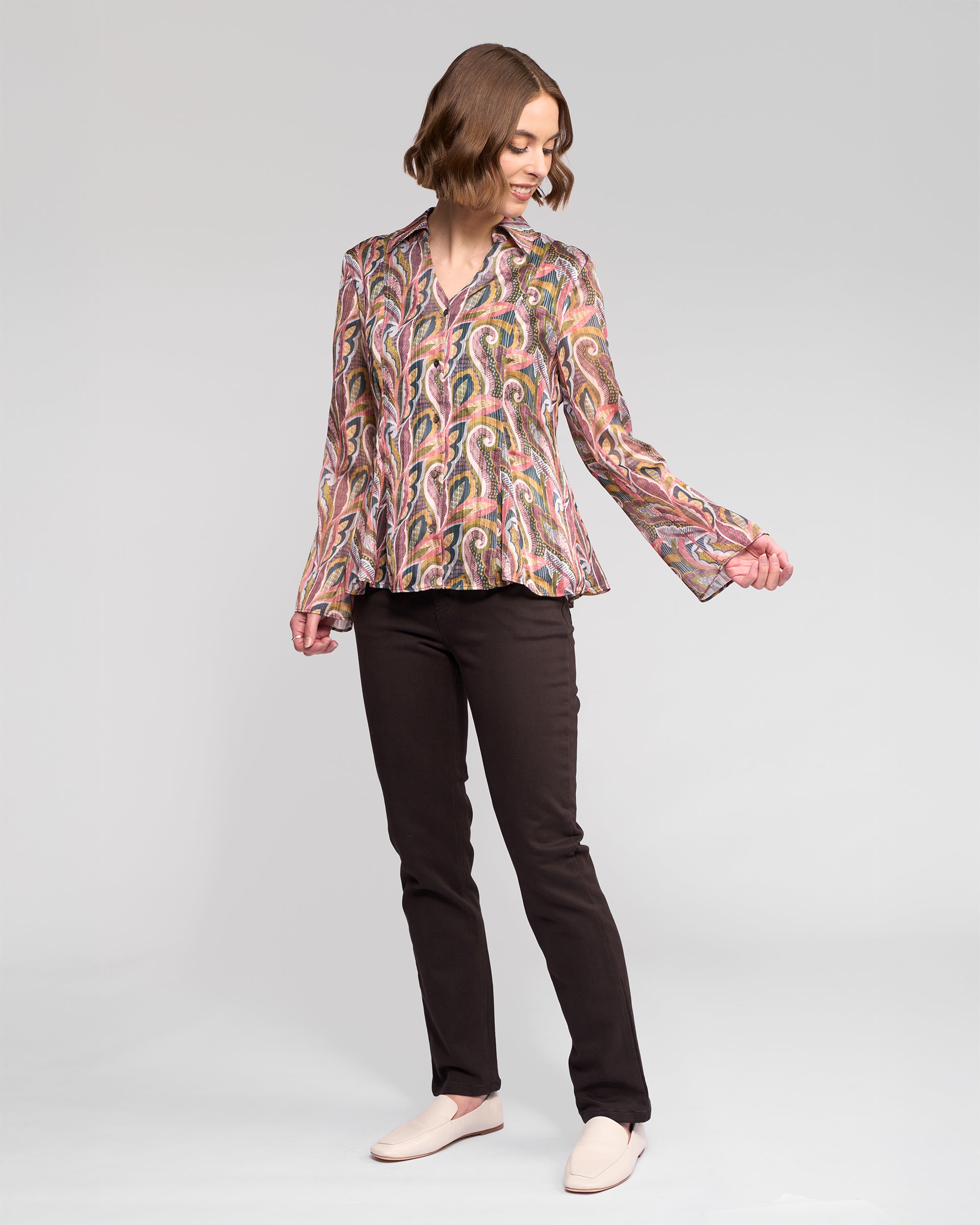 A woman with a short wavy bob wears the Vassalli Valley Shirt in Sable, paired with dark pants and light slip-on shoes, standing and smiling while looking down at her extended hand against a plain background.