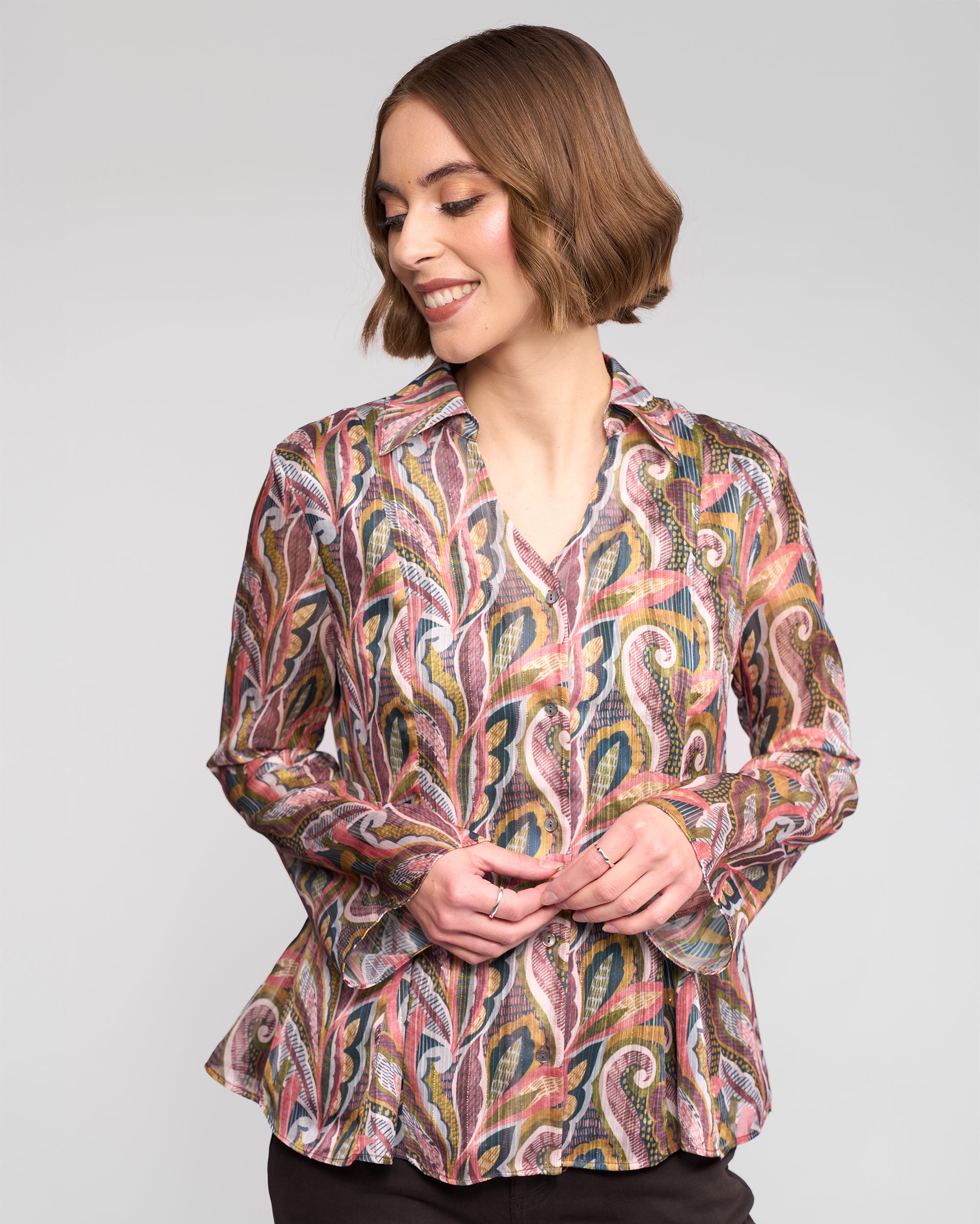 A woman with short brown hair smiles and looks down, wearing the Vassalli Valley Shirt - Sable with colorful, paisley-inspired designs and dark pants, standing against a plain light background.