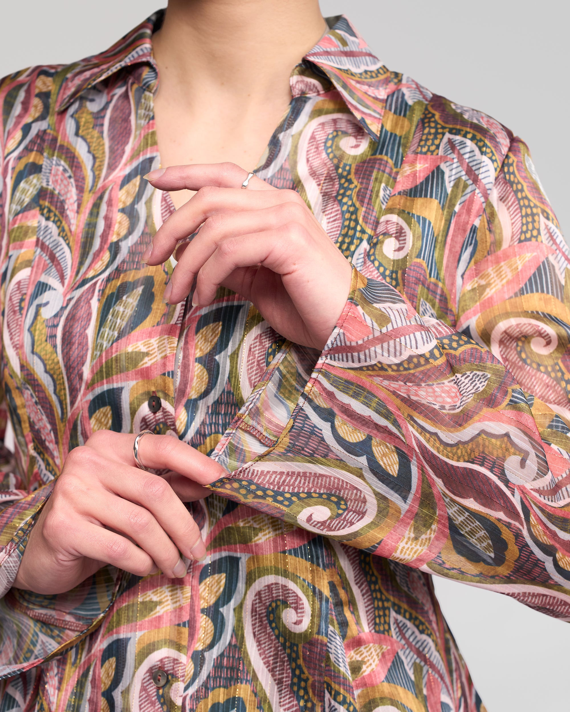 A person in the Vassalli Valley Shirt - Sable, a long-sleeve button-up with colorful paisley-inspired swirling designs, stands against a light background, adjusting their sleeve cuff with their face partially visible.